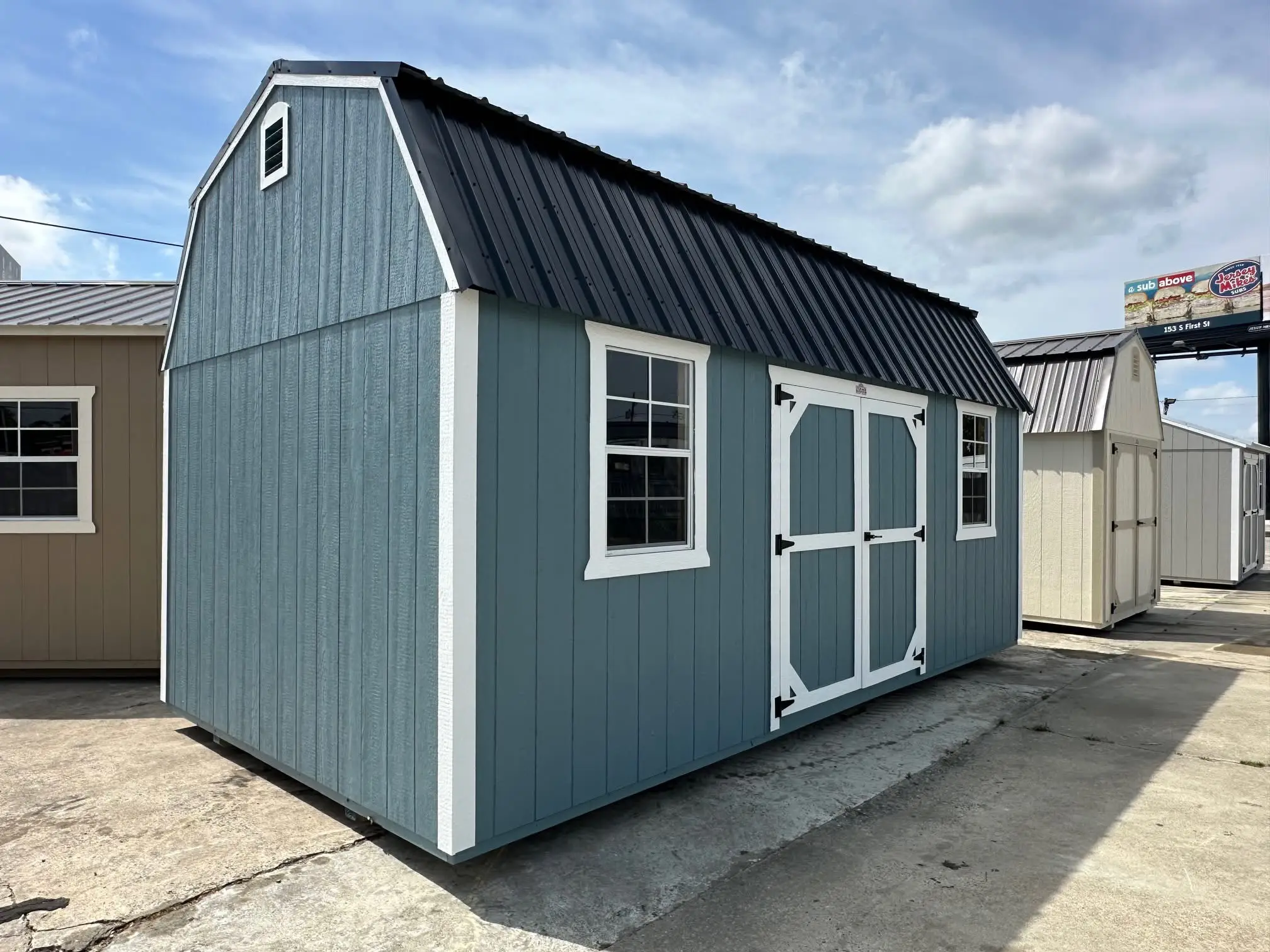 Slate blue lofted barn shed with white trim, double center doors, two windows, and a black metal gambrel roof on a concrete lot in Knoxville, Tennessee.