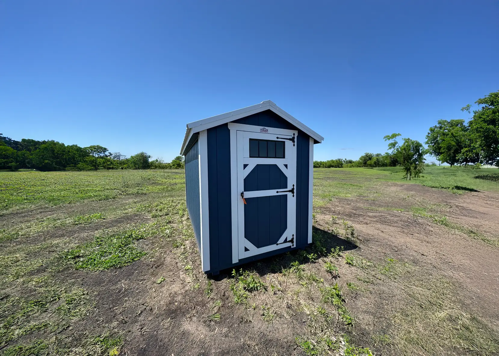 Compact dark blue wooden utility storage shed featuring a single white door with a transom window panel and black T-hinges on a clear grassy lot in The Villages, FL.
