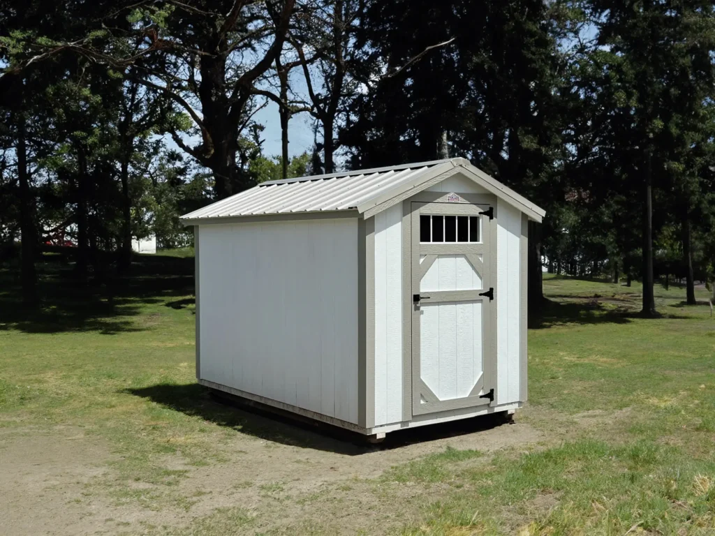 Small white storage shed with grey trim, a silver metal gable roof, and a single front door with a window grid panel sitting on grass in Greenville, South Carolina.