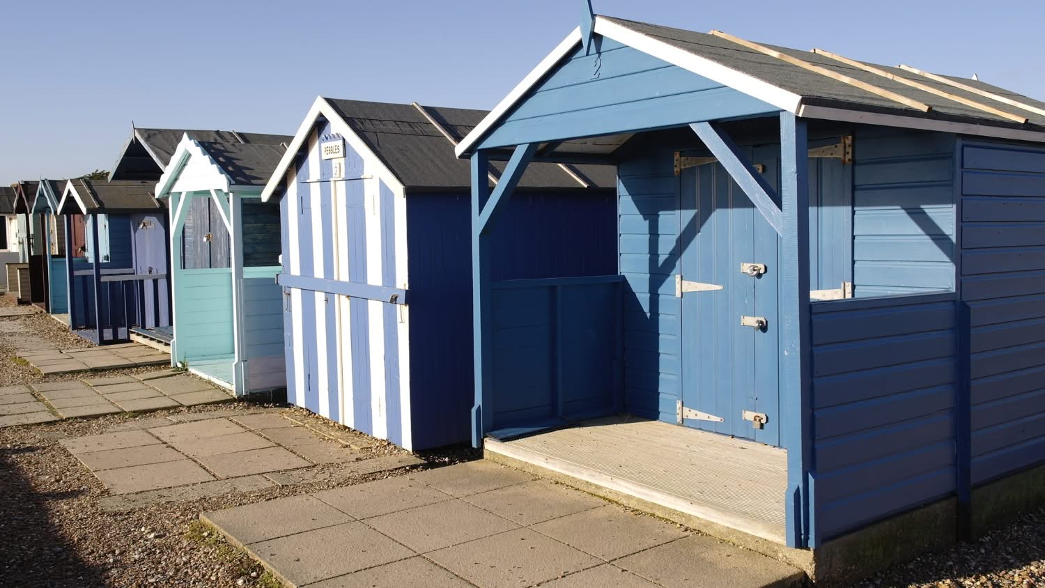 Row of colorful wooden beach hut sheds featuring a prominent blue garden cabana with a front porch overhang, timber siding, and padlocked doors set along a paved walkway in Cape Cod, Massachusetts.
