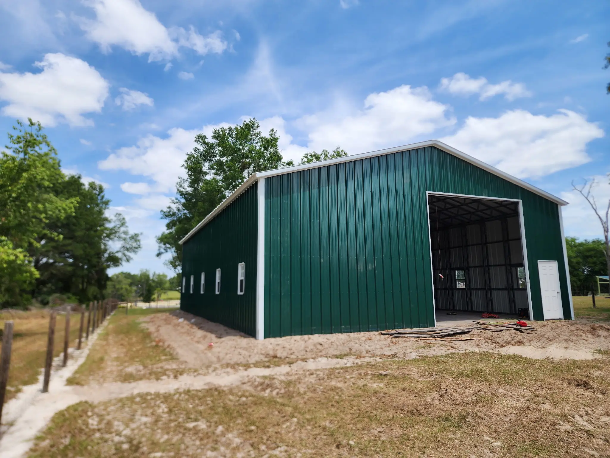 A large dark green metal barn building with a white roof, side windows, open front entry, and side access door on a grassy lot in Dallas, TX.