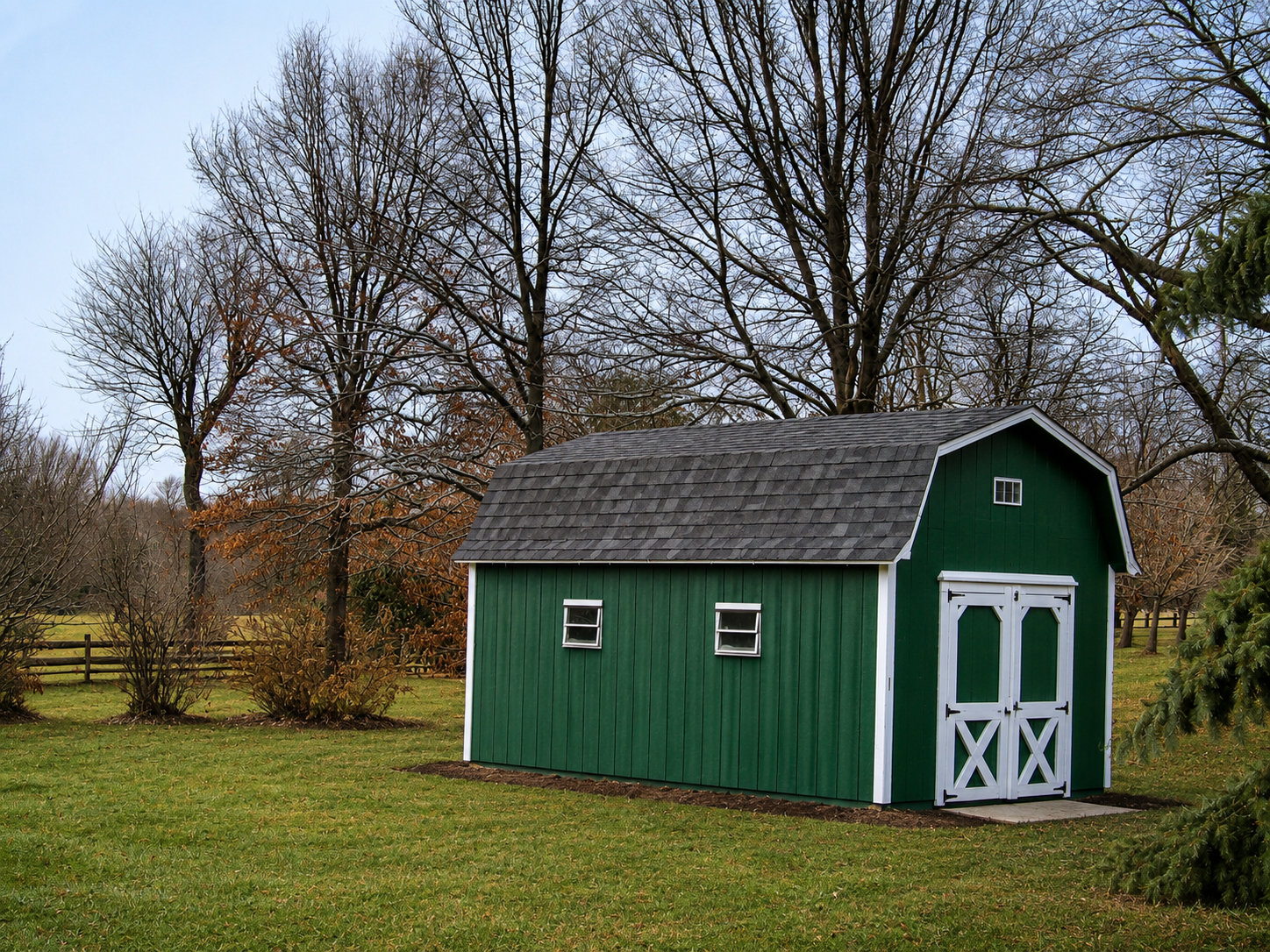 A dark green wooden barn storage shed with white double doors and a dark shingle gambrel roof on a grassy field in Dallas, TX.