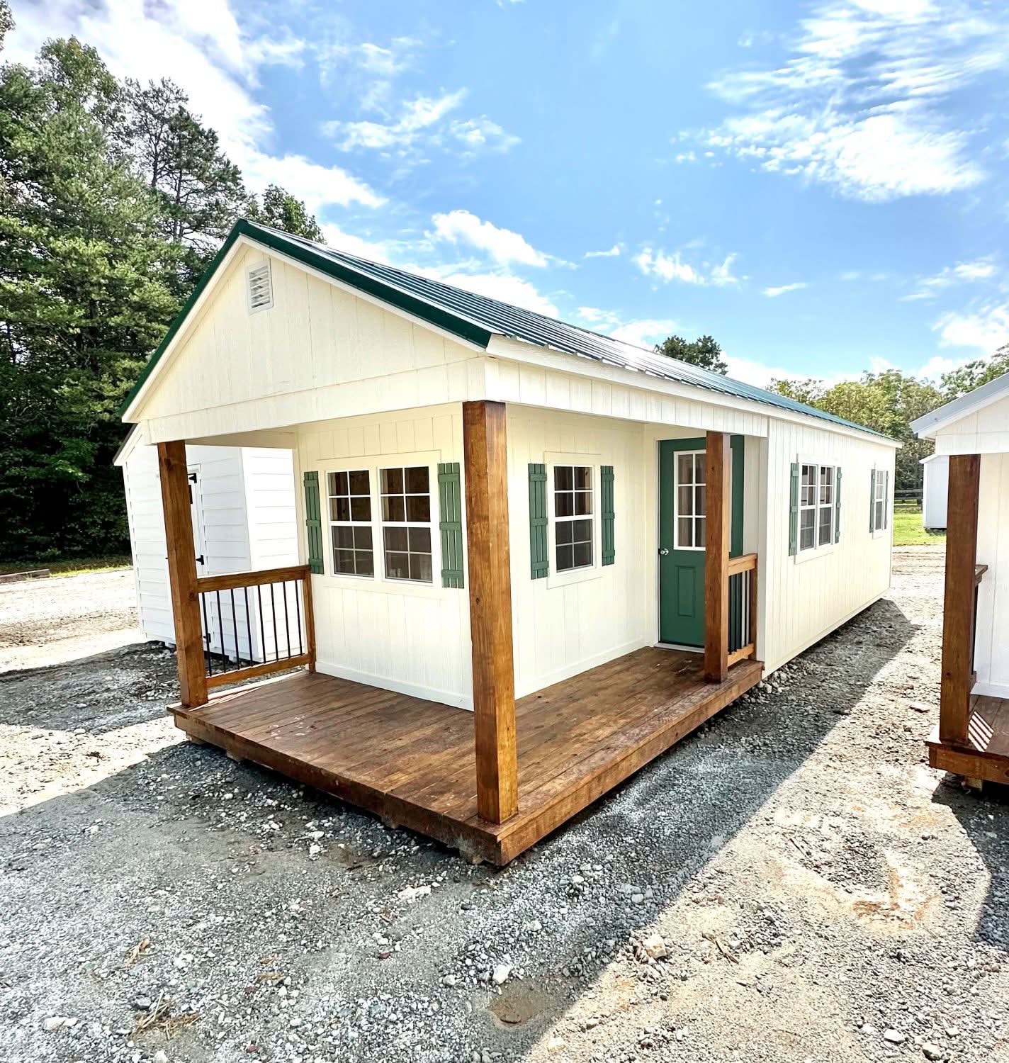 Off-white cabin shed featuring a wrap-around stained wood porch, dark green metal roof, green shutters, and green front door in Boone, North Carolina.