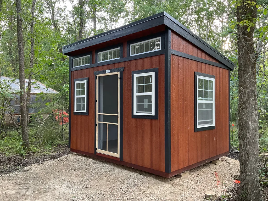 A warm wood-toned modern slant roof studio shed with black trim, a screen front door, and upper transom windows set in a wooded area in Dallas, TX.