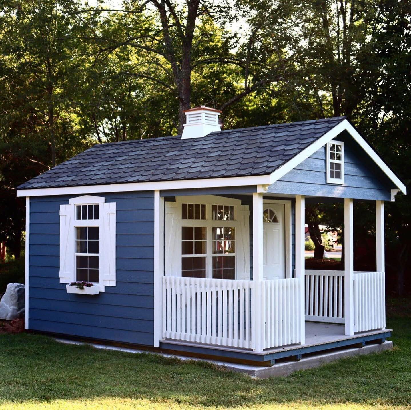 A blue wooden cabin shed featuring a covered front porch with white railings, window shutters, and a rooftop cupola in Dallas, TX.