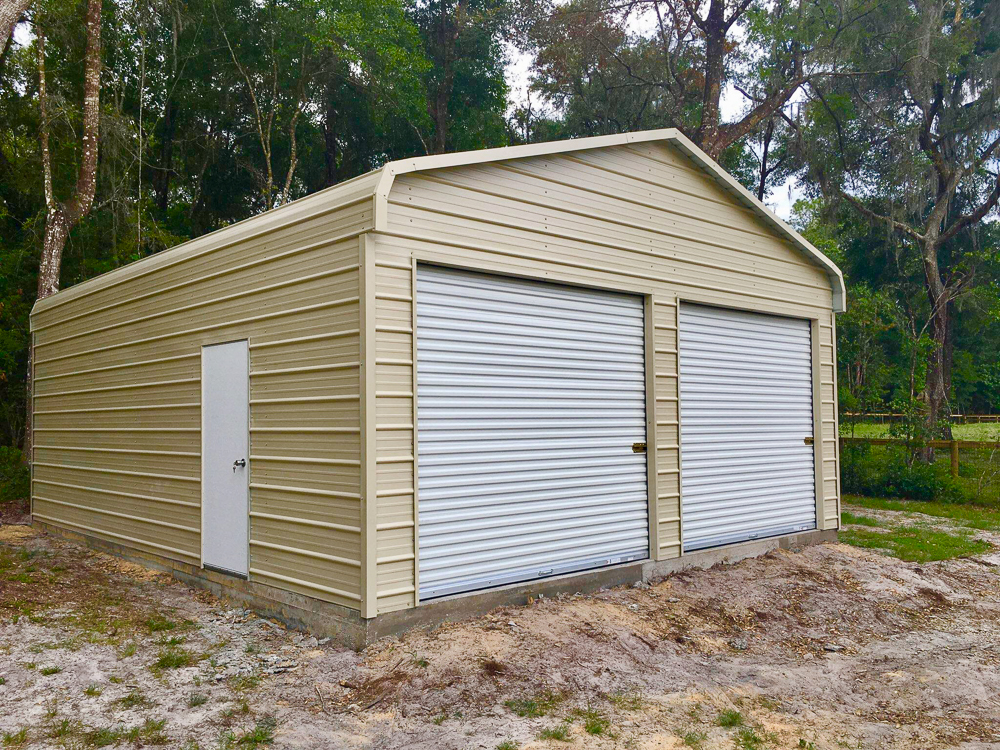 A beige two car metal garage with dual white roll-up doors, a side walk-in door, and a concrete foundation installed on a wooded lot in The Villages, FL.