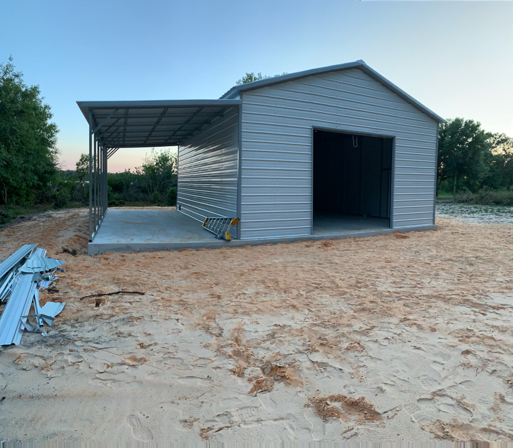 Newly completed light gray custom metal garage installation featuring an open bay door and attached side lean-to carport on a concrete pad in Inverness, FL.