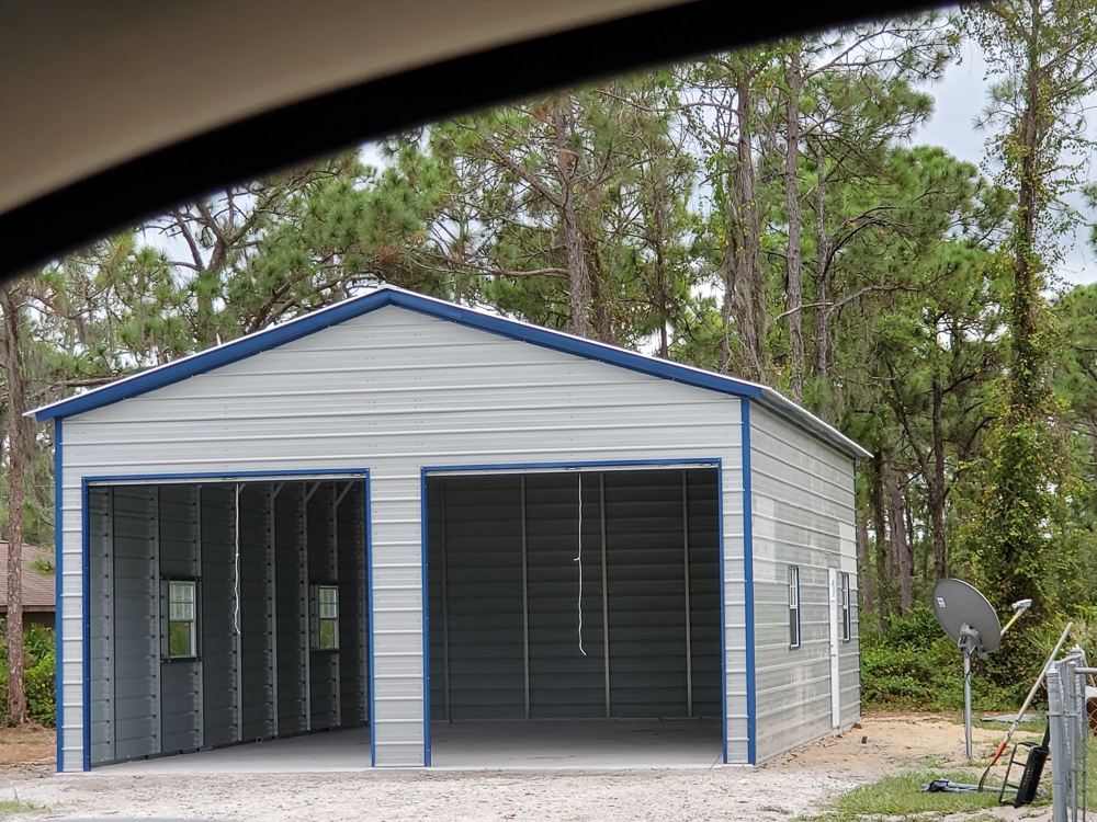 Light gray two door metal garage with blue trim, dual open bay doors, and side windows surrounded by pine trees in Dunnellon, FL.