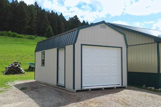 Beige lofted barn garage shed featuring dark green trim, a roll-up garage door, white side walk-in door, and a metal gambrel roof in Missoula, Montana.