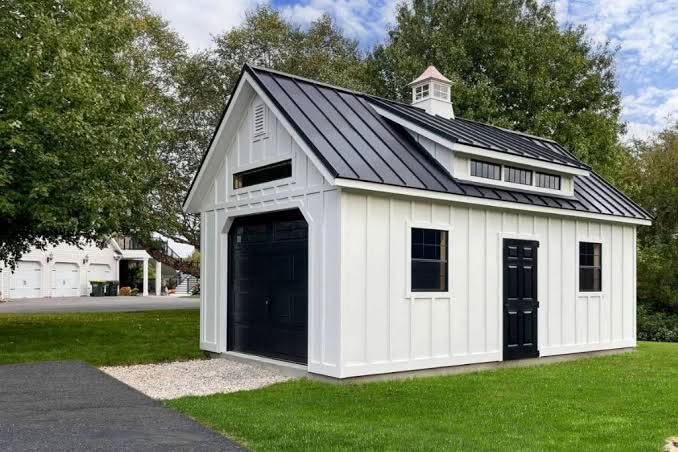 White board and batten modern farmhouse shed with a black metal roof, cupola, and black garage door set on a green lawn in Lancaster, Pennsylvania.