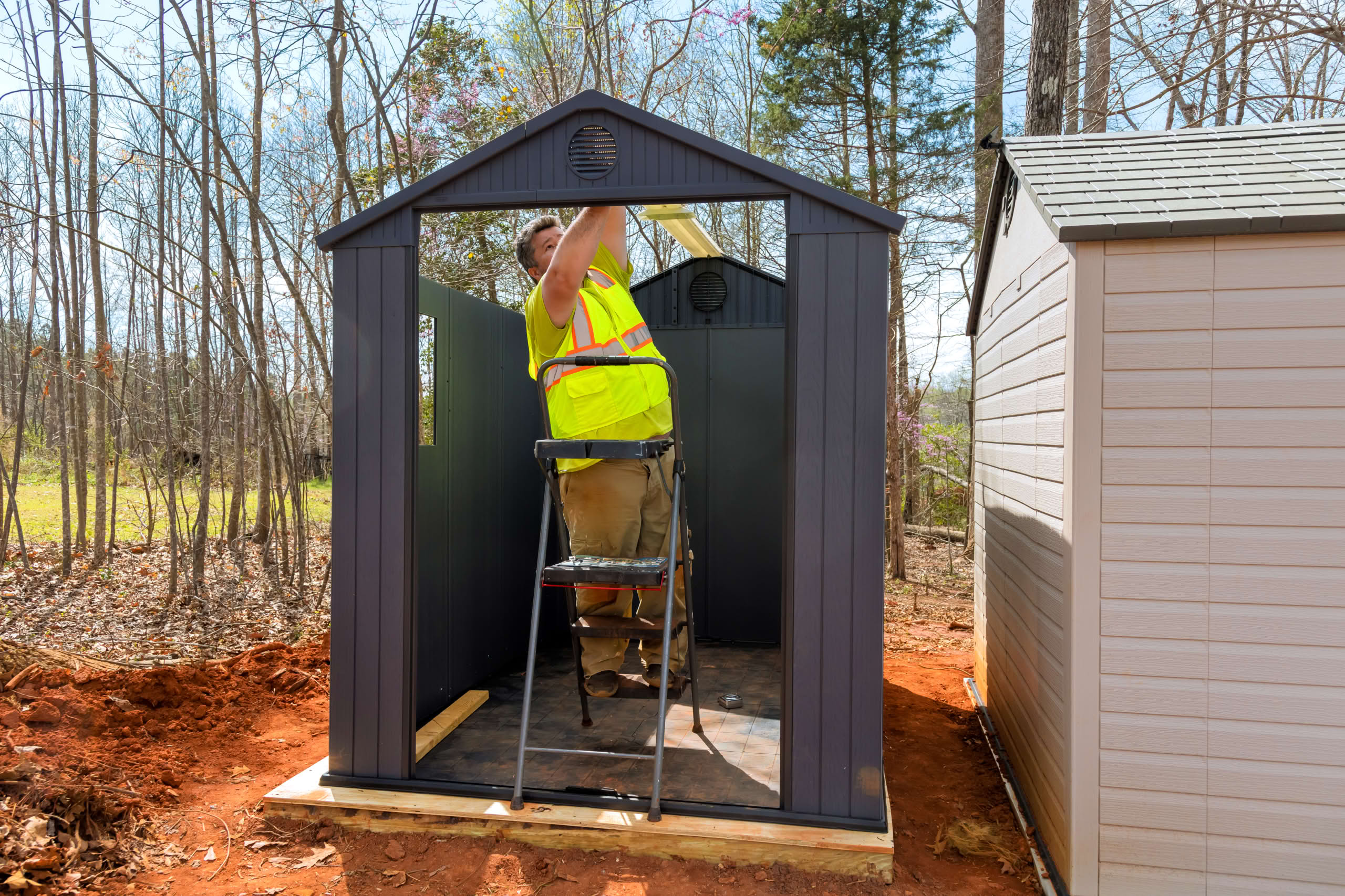 Worker assembling storage shed in clearing surrounded by trees during daylight hours