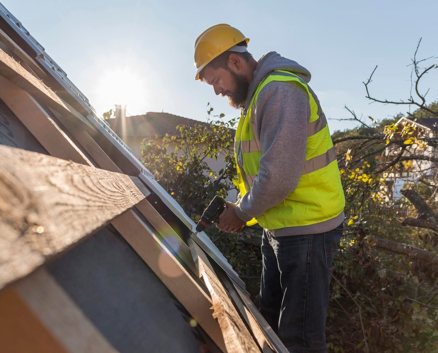 A professional contractor wearing a yellow hard hat and safety vest using a power drill to install roofing boards on a shed in Dallas, TX.