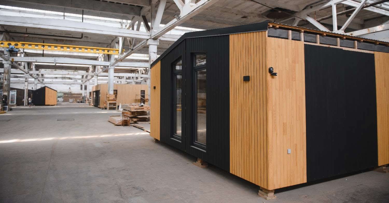 A modern prefabricated modular studio office with black and natural wood paneling under construction inside a large manufacturing warehouse in Dallas, TX.