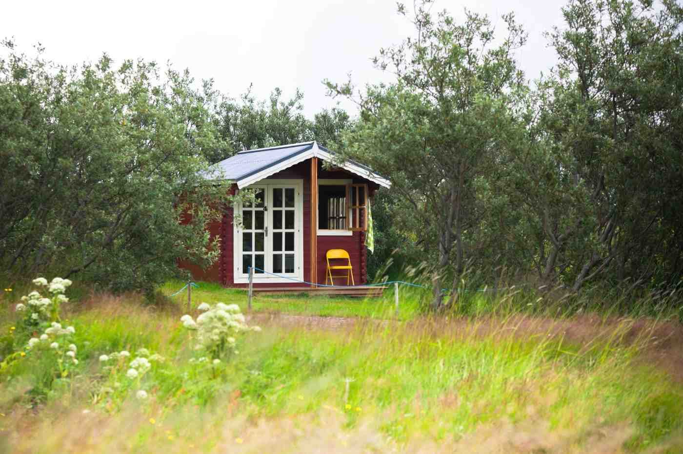 Small red wood cabin shed studio with white glass french doors, a small side porch, and a yellow chair nestled in a lush green field in Boise, Idaho.