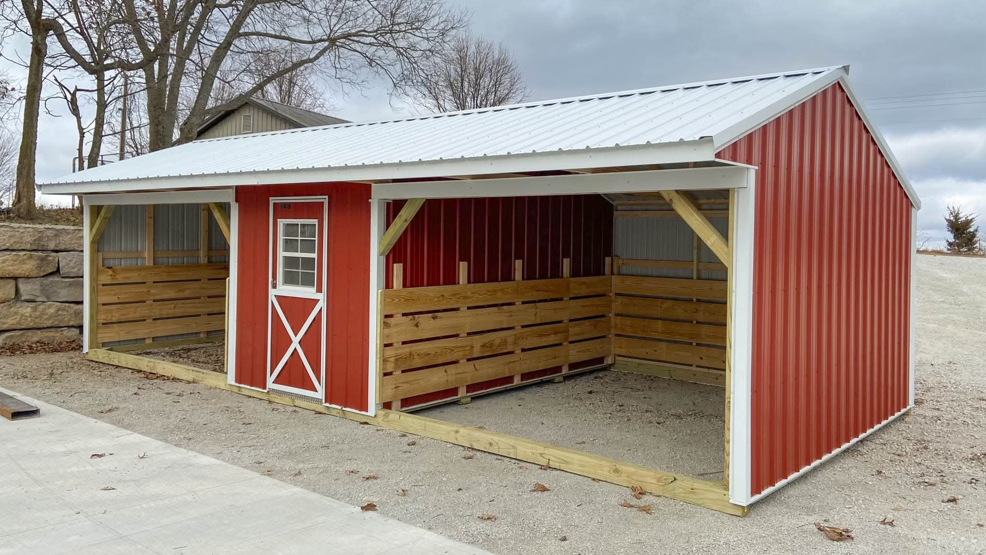 A red metal loafing shed with two open horse shelter bays flanking a central tack room with a white cross-buck door in Dallas, TX.