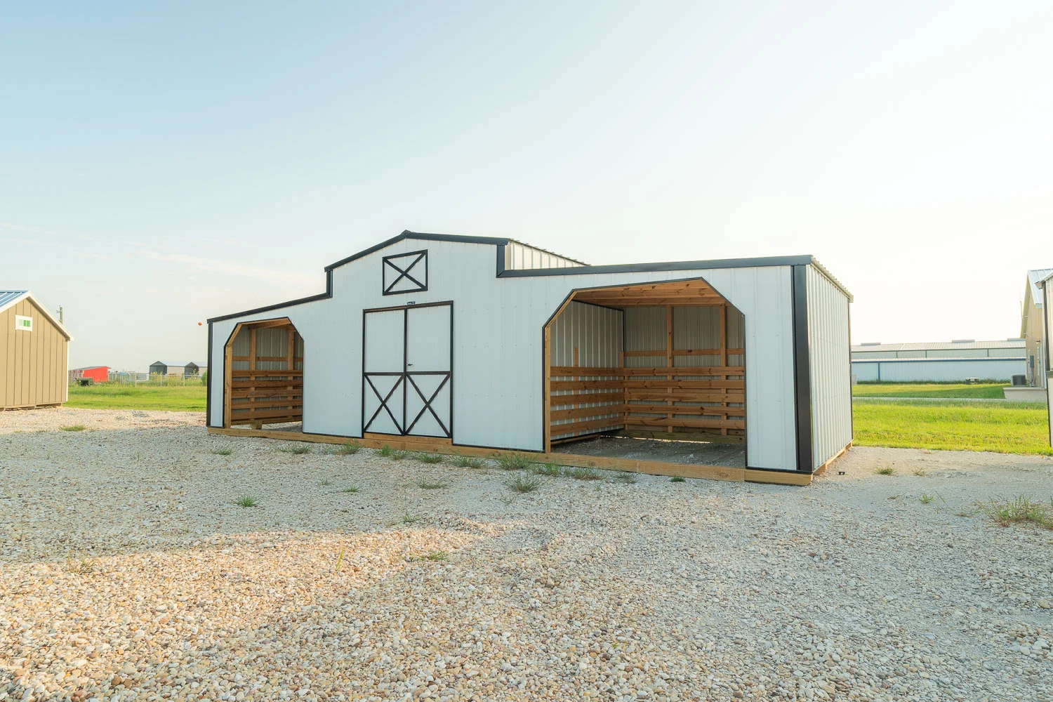 White metal horse barn shed featuring black trim, a center enclosed tack room with double doors, and two open-front side run-in lean-to shelters set on gravel in Aiken, South Carolina.