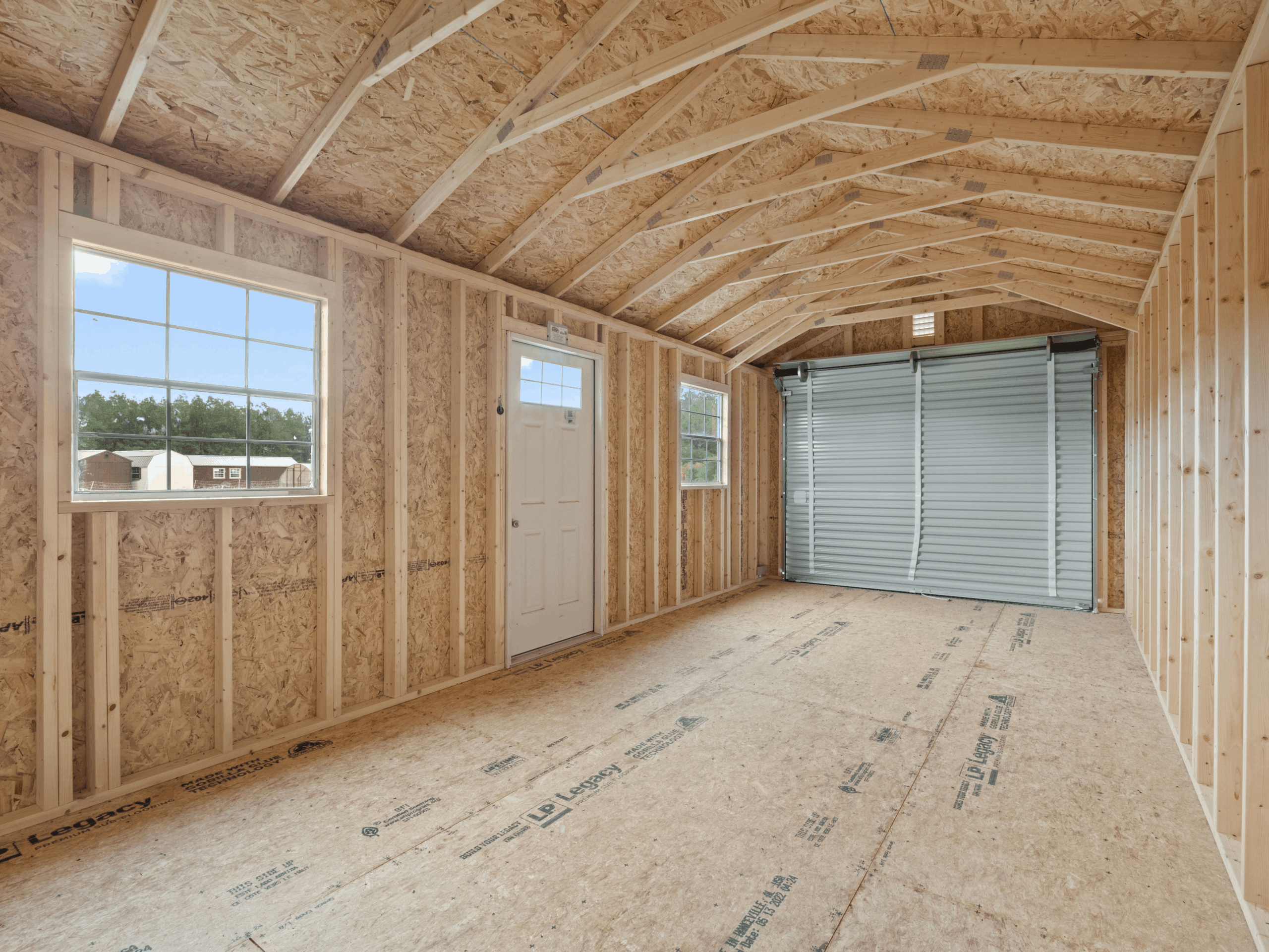Interior view of an spacious, unfinished 12x24 garage storage shed featuring LP Legacy subflooring, wooden wall framing, grid windows, and a roll-up garage door in Clermont, FL.