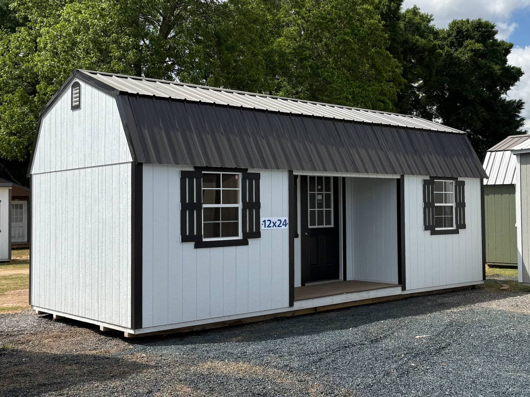 White 12x24 lofted barn cabin shed with black trim, dark metal gambrel roof, recessed side porch entry, and black window shutters in Ocala, FL.