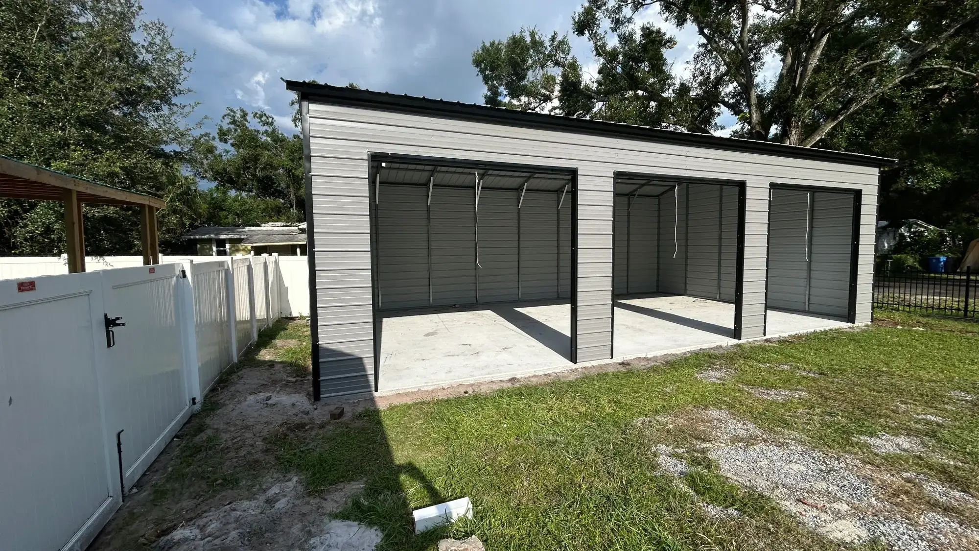 Newly installed light grey 3 car metal carport with open frame bay doors and concrete slab foundation on a residential yard in Orlando, FL.