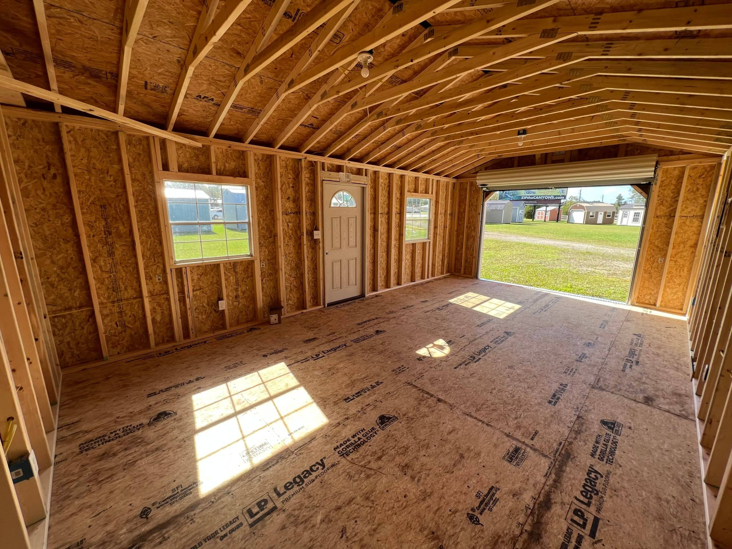 Interior view of a wide wooden garage shed showing exposed wall framing, LP Legacy subflooring, side door, windows, and an open roll-up garage door in Crystal River, FL.