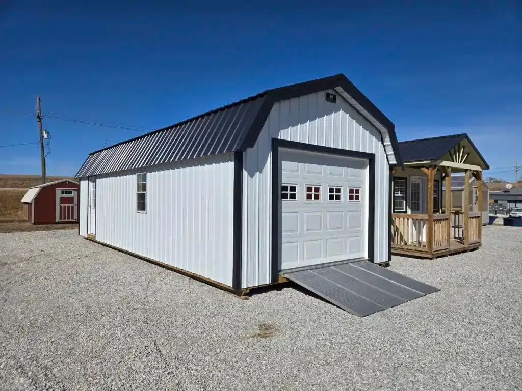 Exterior view of a white high-barn garage shed with black trim, a panel garage door, entry ramp, side door, and metal roof on a gravel lot in Inverness, FL.
