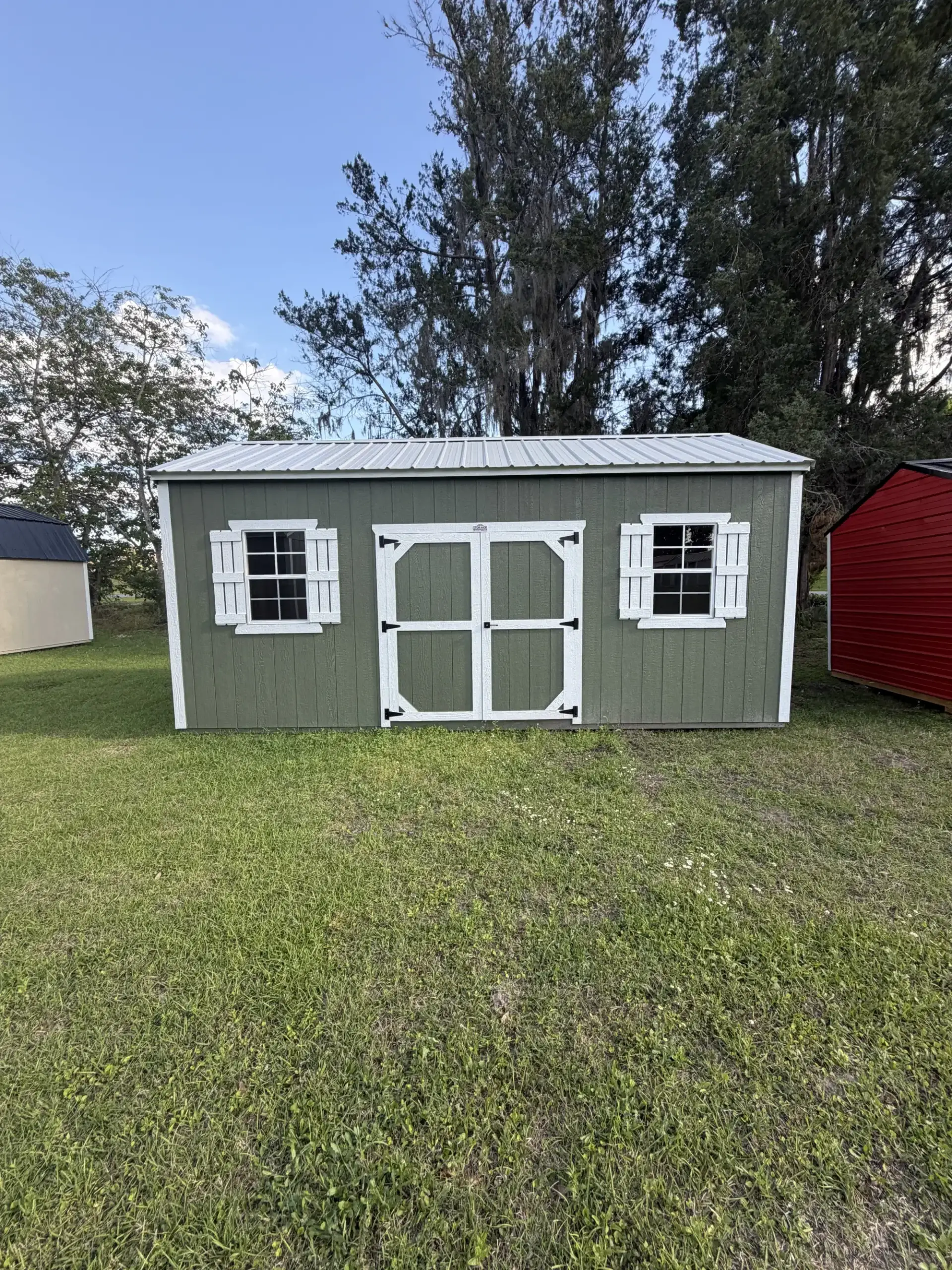 Olive green custom garden storage shed featuring white trim, decorative window shutters, double doors, and a silver metal roof on a grassy field in Inverness, FL.