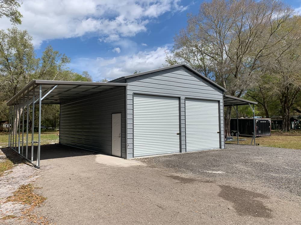 Large gray custom metal garage featuring dual roll-up doors, a side walk-in door, and open lean-to carport extensions on a dirt driveway in Crystal River, FL.