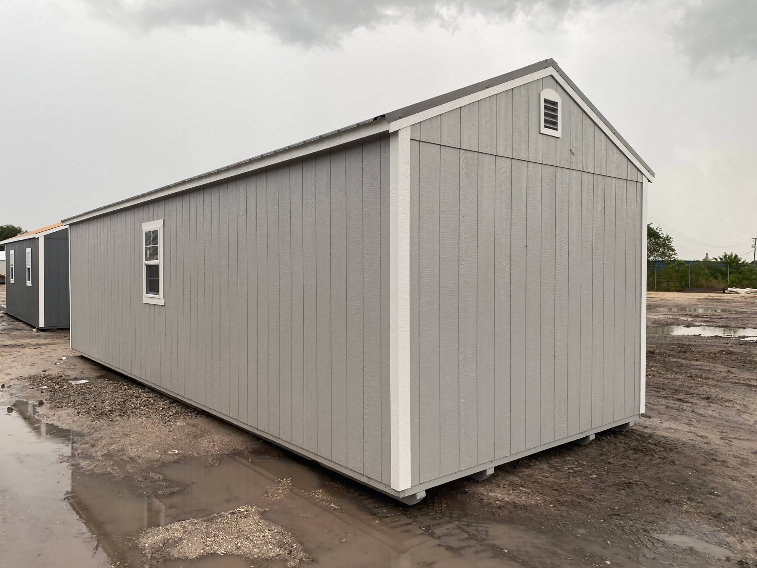 Exterior rear view of a long light-gray custom outdoor shed with white trim, gable roof, and side window on a dirt lot in Crystal River, FL.