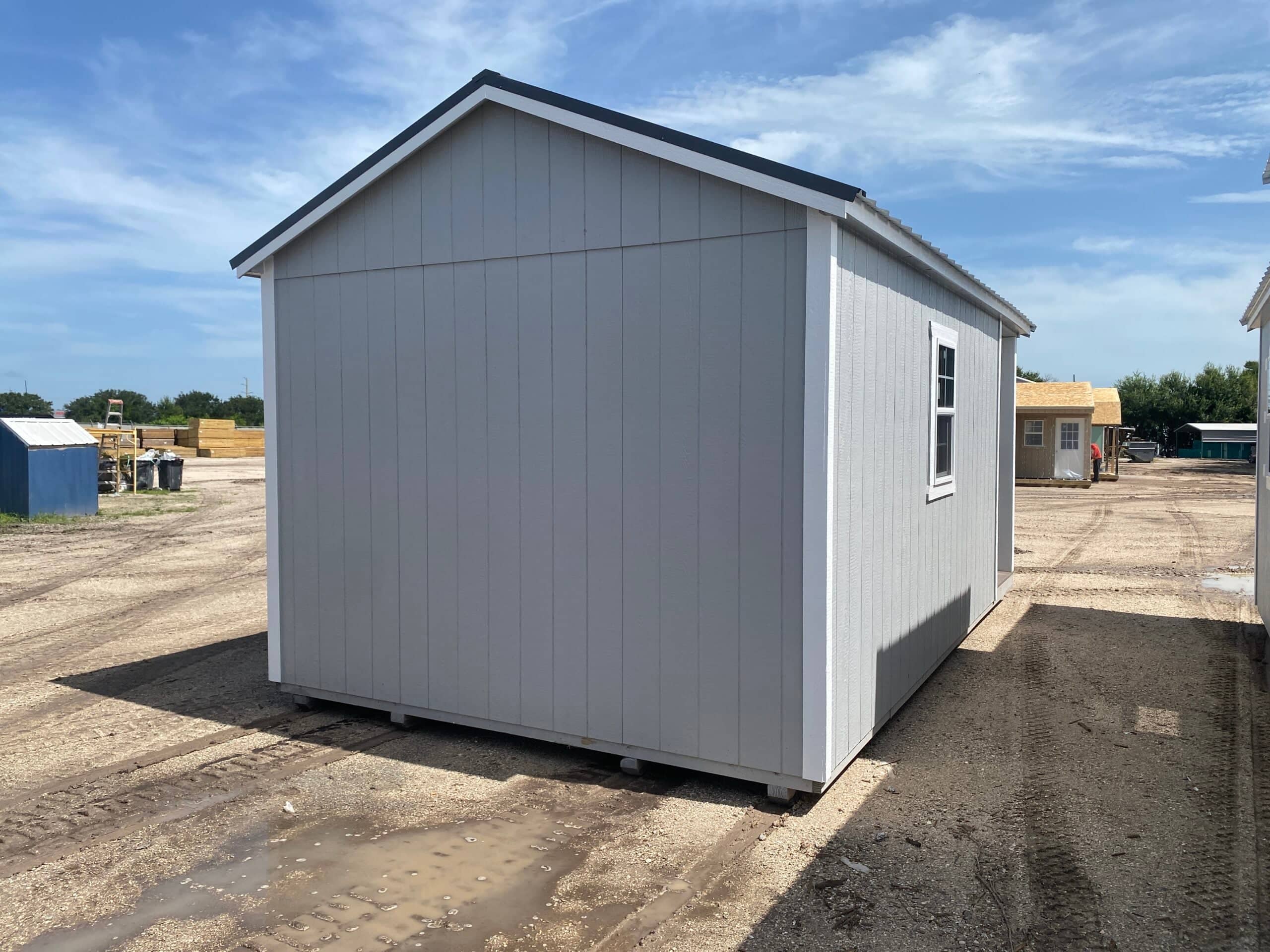 Rear corner view of a light gray custom outdoor storage shed featuring white trim, a gable roof, and a single side window on a sand lot in Ocala, FL.