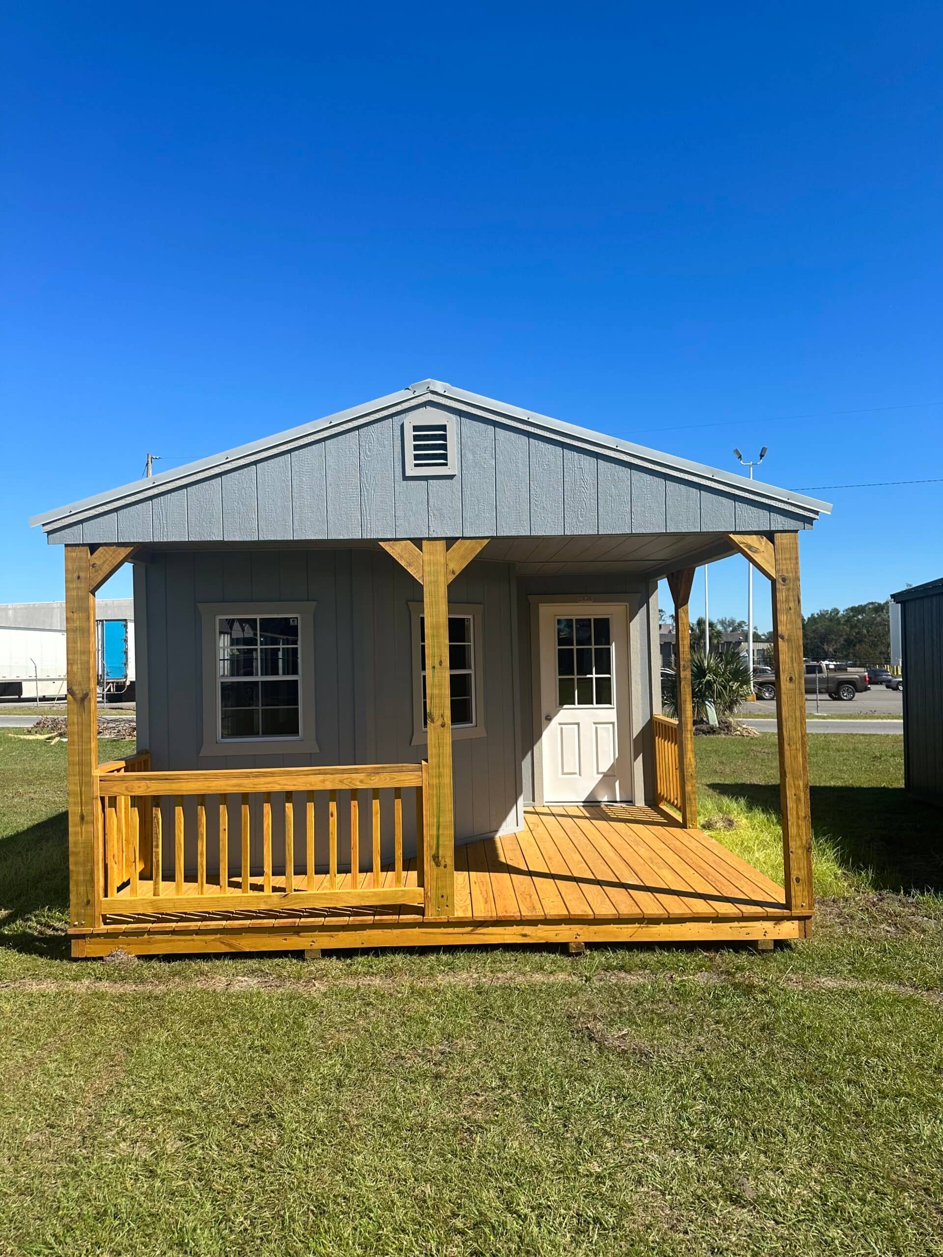 Front view of light grey custom wooden cabins featuring a wooden porch deck, railing, timber posts, two grid windows, and a white front door in Gainesville, FL.