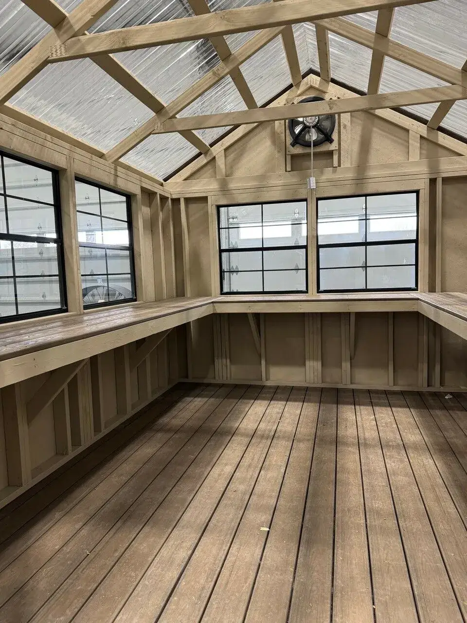 Interior view of a custom wooden greenhouse featuring U-shaped potting benches, translucent roof panels, black grid windows, and an exhaust fan in Dunnellon, FL.