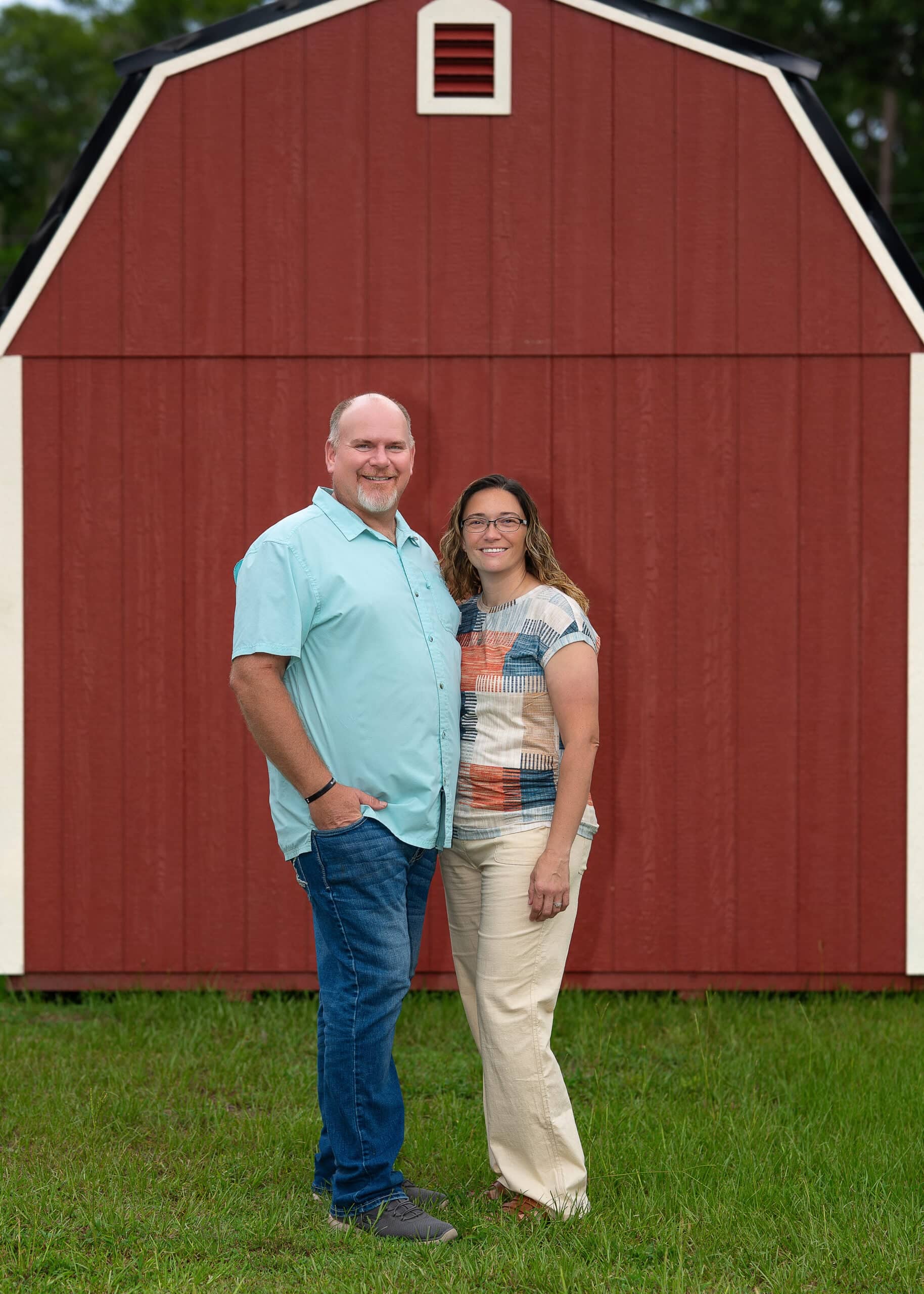 A man and woman standing on a grass lawn in front of a red gambrel barn-style storage building, provided by custom wooden sheds specialists in Leesburg, FL.