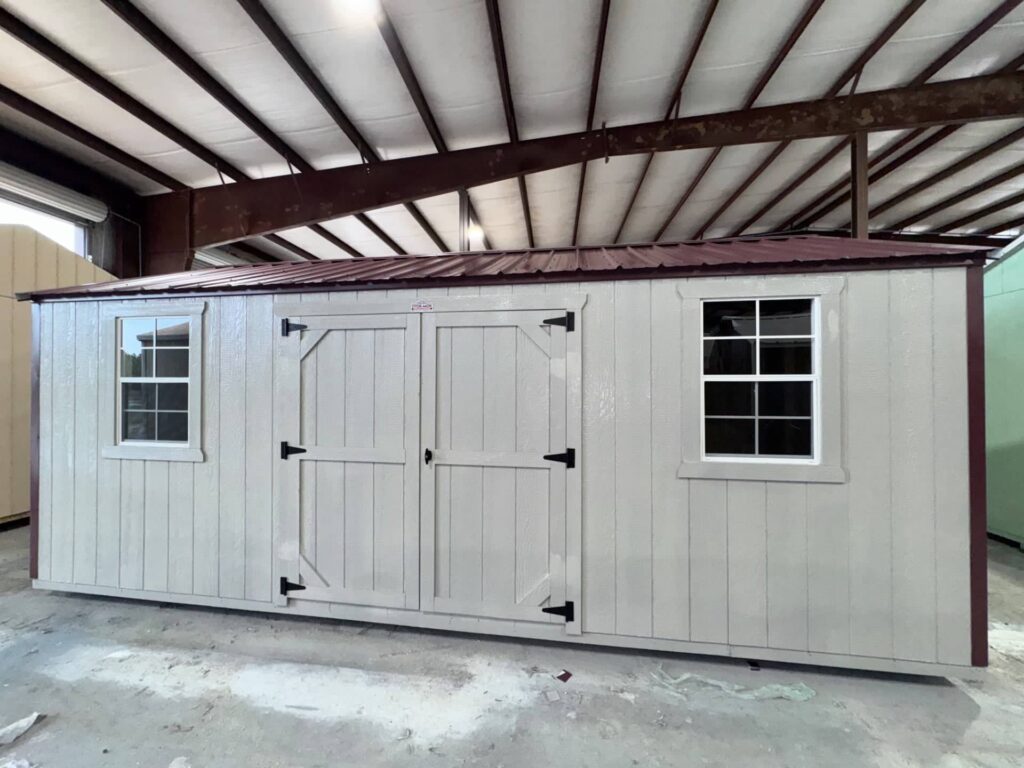 Light grey custom wooden utility shed featuring dark red roof trim, double entry doors, and two grid windows inside a manufacturing facility in Dunnellon, FL.
