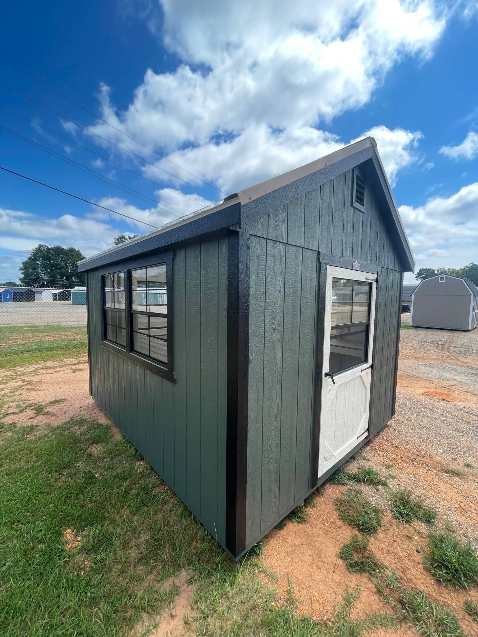 Dark green utility storage shed with black trim, double side windows, and a white entry door set outdoors under a blue sky in Ocala, FL.