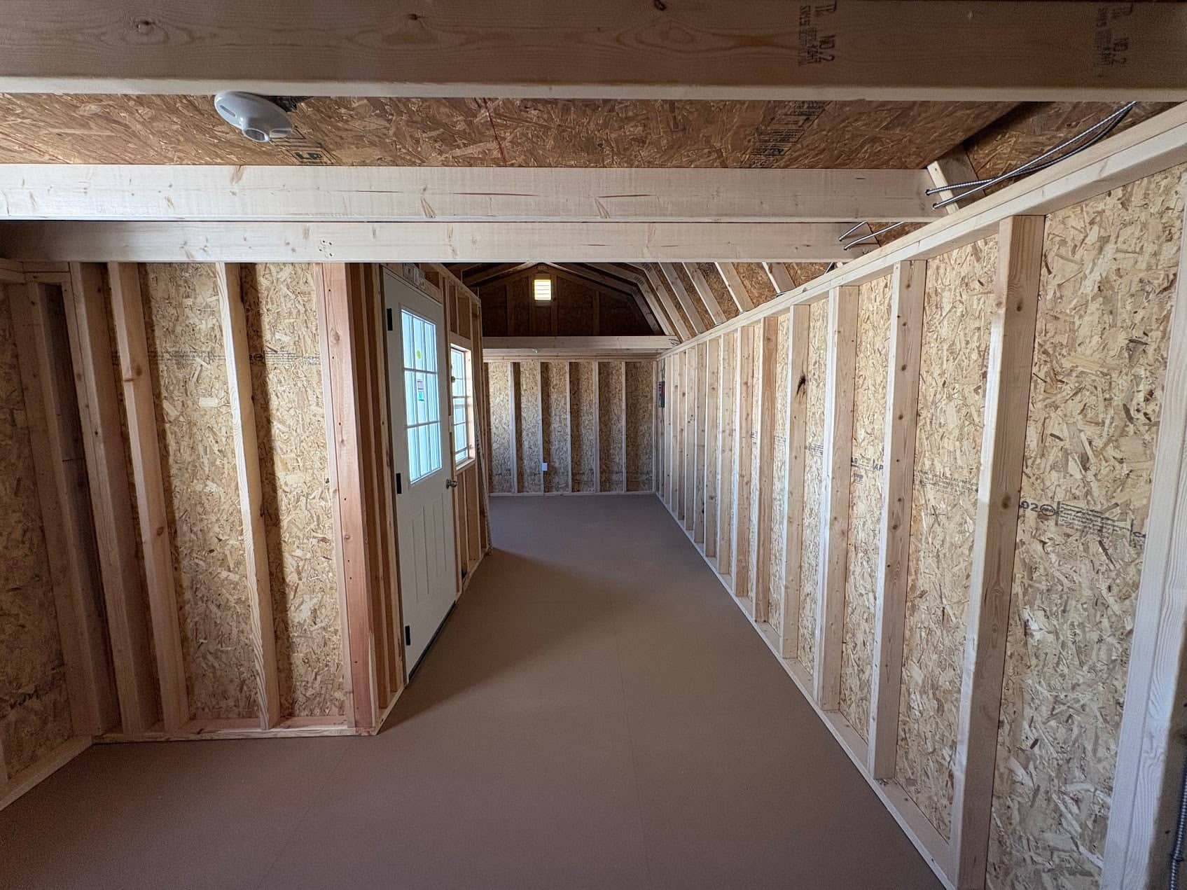Interior view of an unfinished wooden lofted barn cabin showing wall studs, OSB sheath paneling, smooth flooring, white entry door with grid windows, and overhead loft framing in Dunnellon, FL.