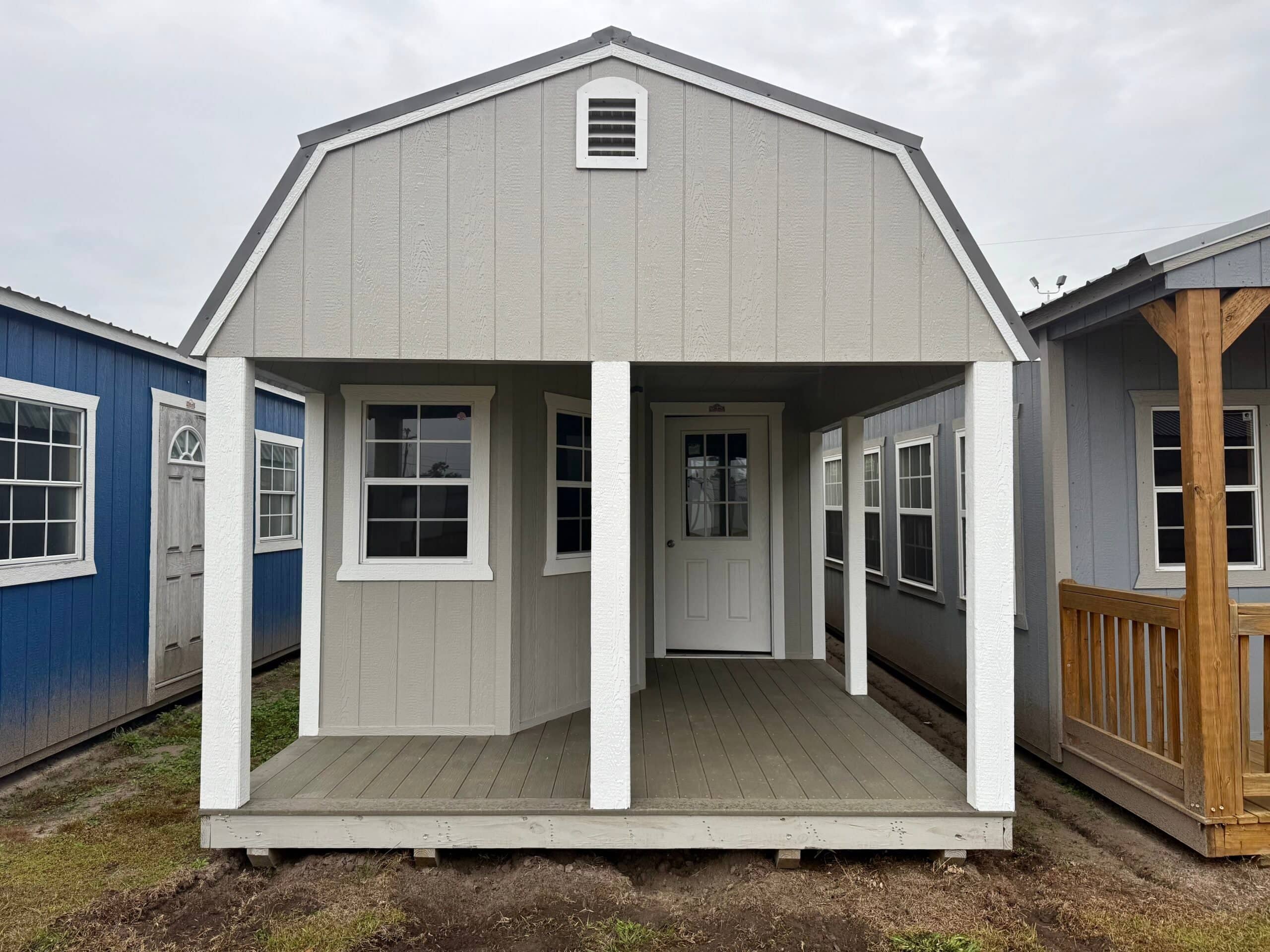 Light grey wooden lofted barn cabin shed featuring white accent trim, a covered front porch, white support posts, a central glass panel entry door, and side grid windows on a dirt lot in Bronson, FL.