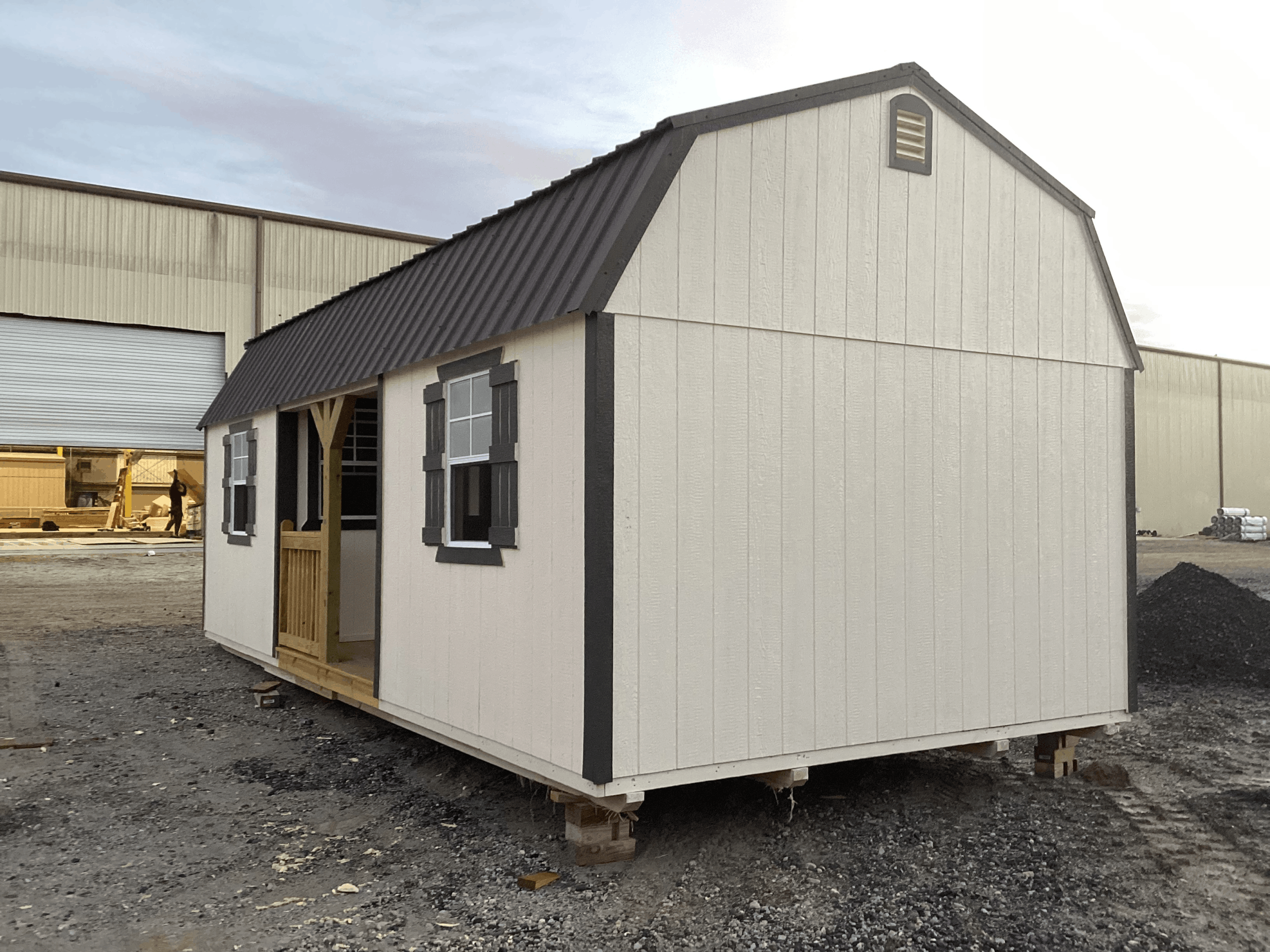 White wooden lofted barn cabin shed featuring dark trim and shutters, grid window, small side porch entry deck with wooden railing, and a dark metal gambrel roof in Brooksville, FL.