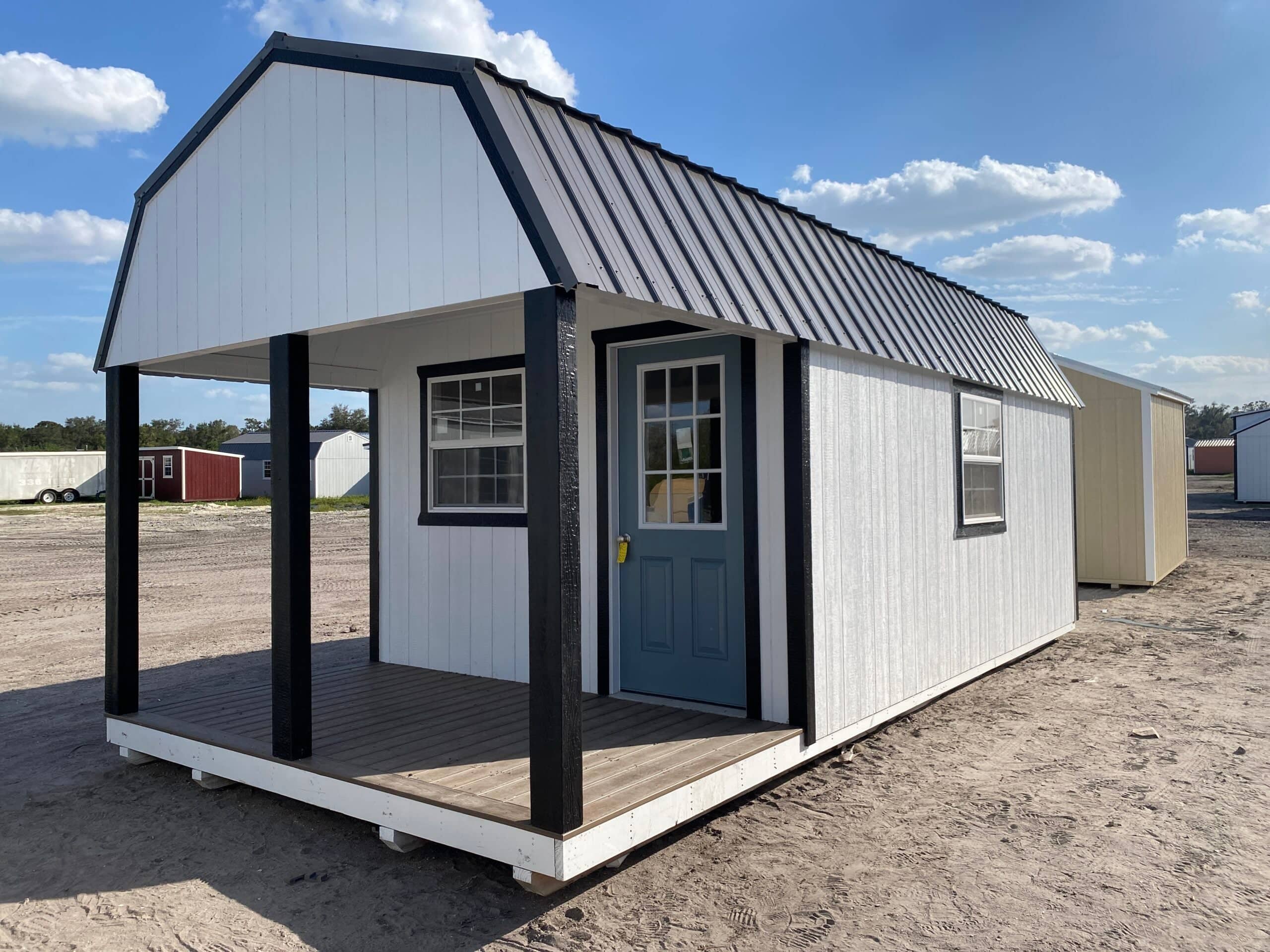 White wooden lofted barn cabin shed featuring bold black porch posts and trim, a blue glass-paneled entry door, covered porch deck, side grid window, and dark metal gambrel roof on a gravel lot in Brooksville, FL.