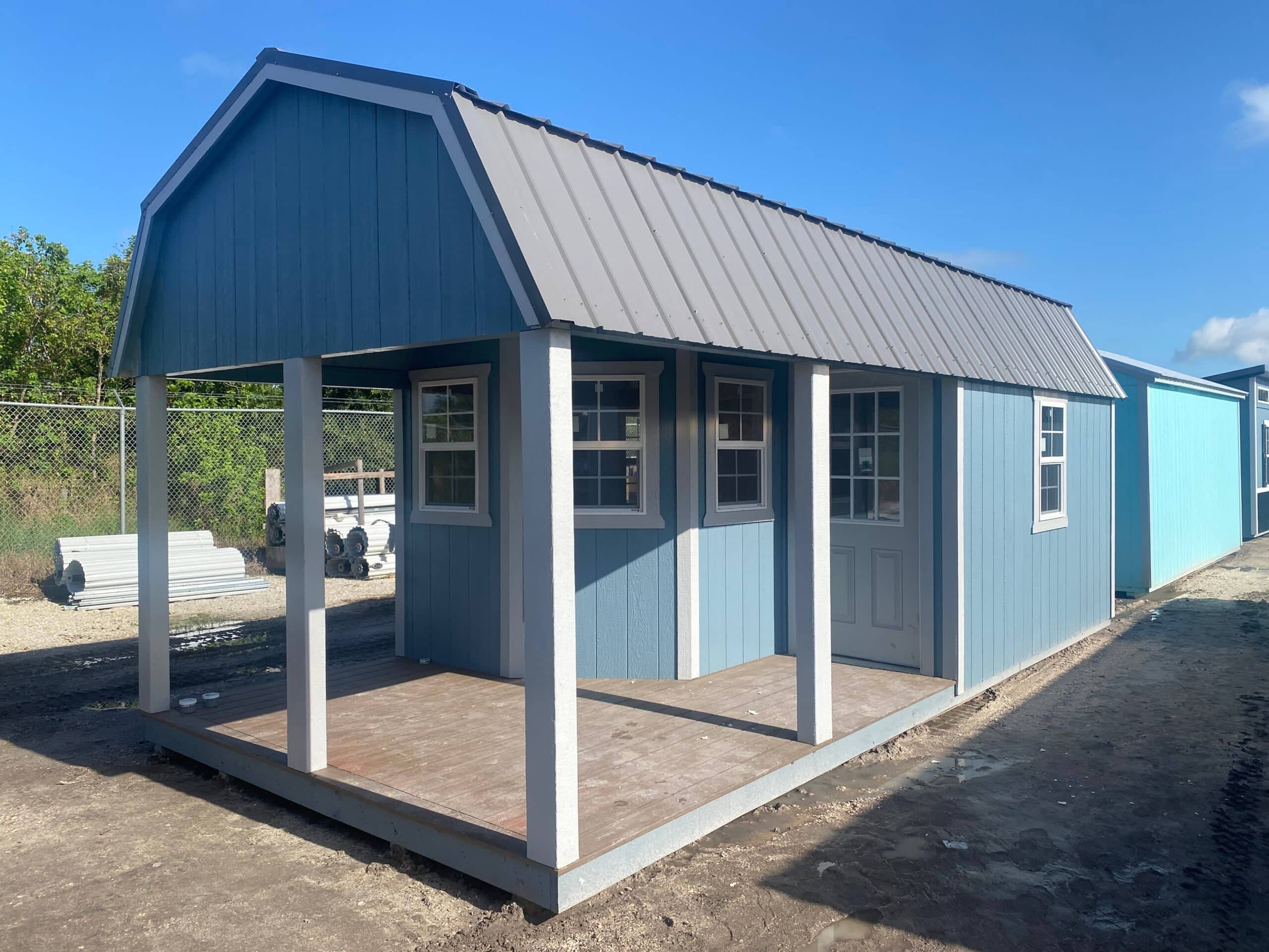 Slate blue wooden lofted barn cabin shed with white trim, side porch decking, white posts, grid windows, glass panel door, and grey metal roof in Crystal River, FL.