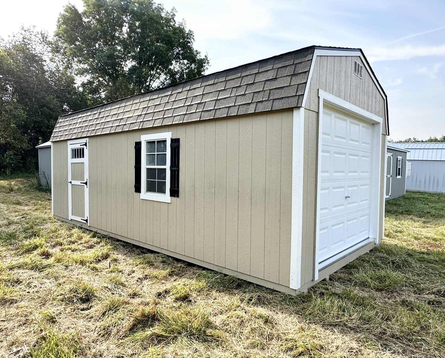 Exterior side view of a beige high-barn garage shed featuring white trim, brown shingle roof, white sectional garage door, side door, and black shutters in Alachua, FL.