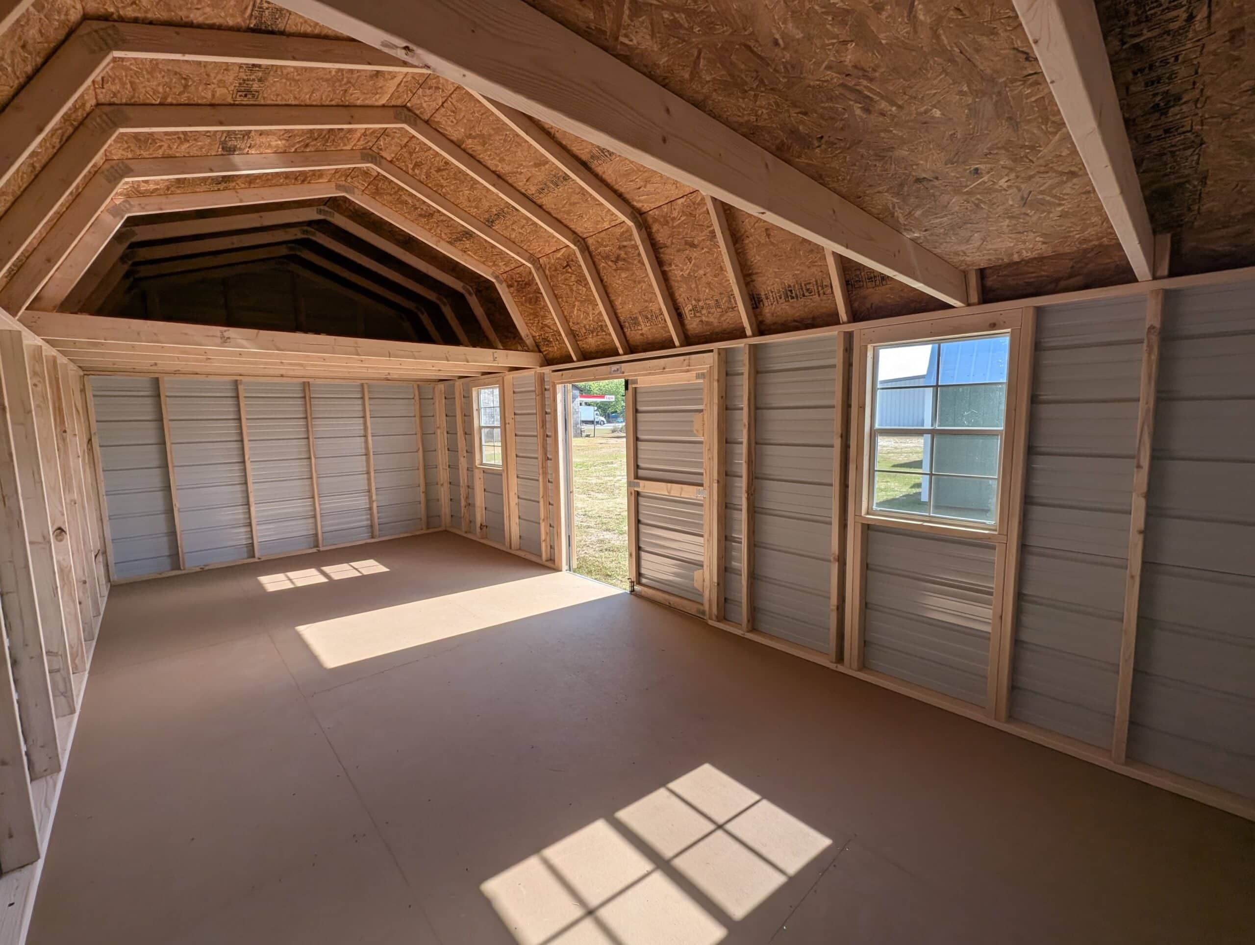 Interior view of an unfinished wooden lofted barn shed showing exposed framing, overhead loft storage area, side window, open door, and smooth floor in Wildwood, FL.