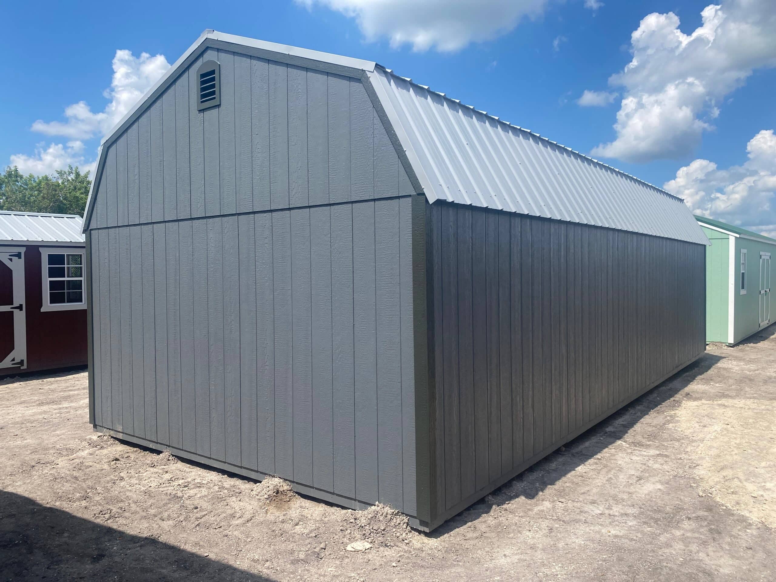 Rear exterior view of a long, dark grey high-barn storage shed with a silver metal roof resting on a sand lot under a blue sky in Gainesville, FL.