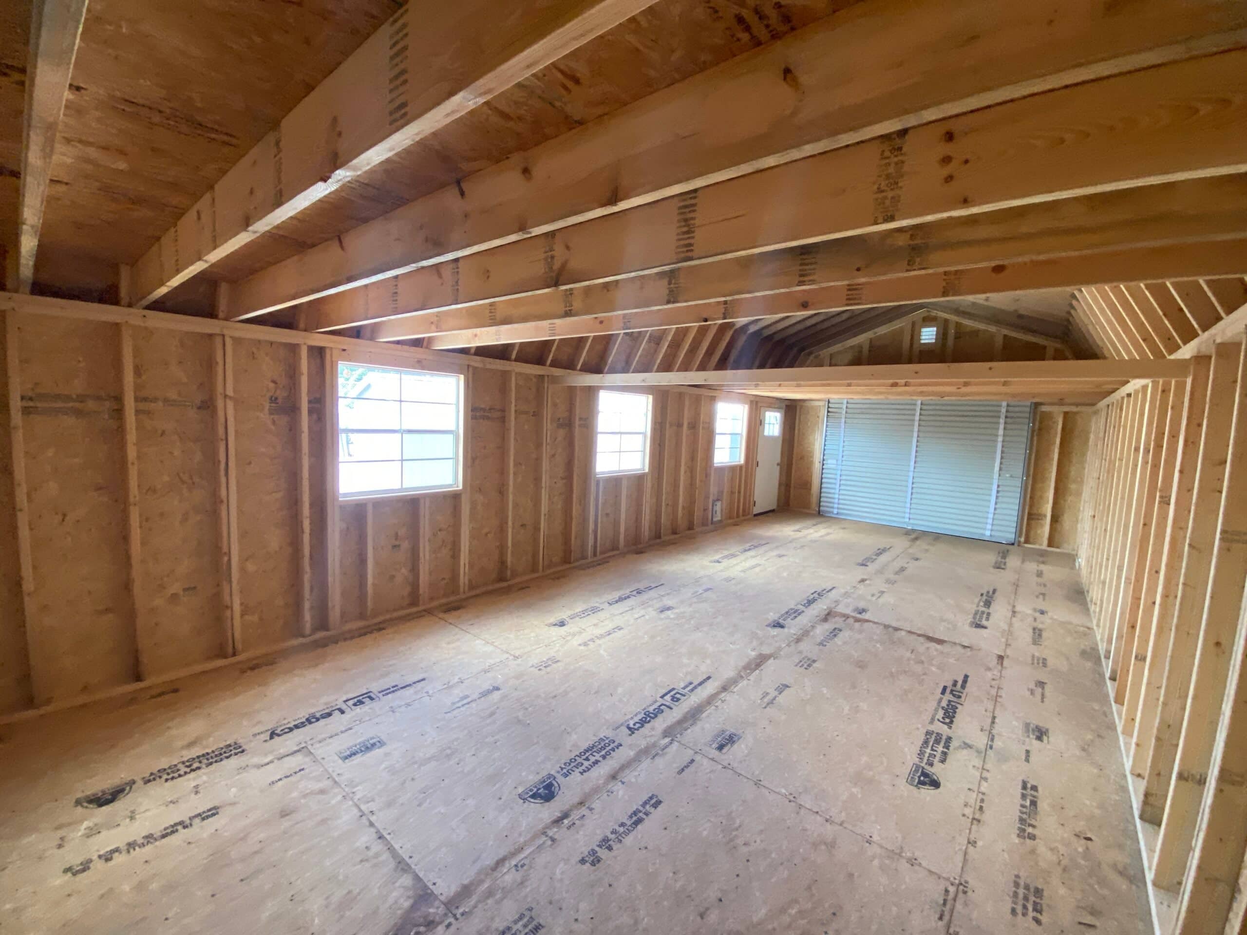 Interior view of a wide lofted garage storage shed showing exposed wood framing, LP Legacy floor panels, loft joists overhead, grid windows, and a far roll-up door in Spring Hill, FL.