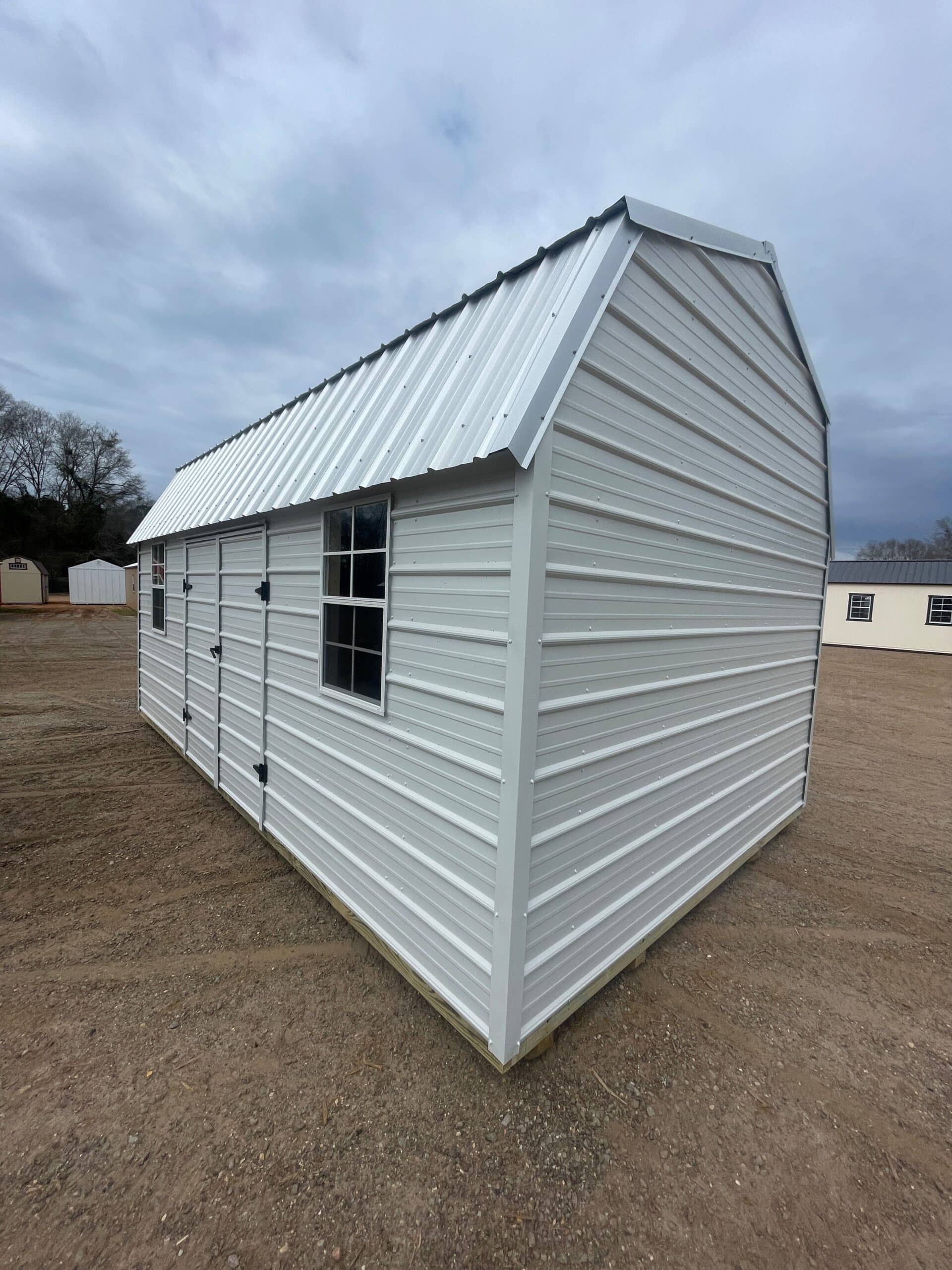 Exterior corner view of white metal barn sheds featuring a silver gambrel metal roof, double doors with black hinges, and grid windows on a gravel lot in Silver Springs, FL.