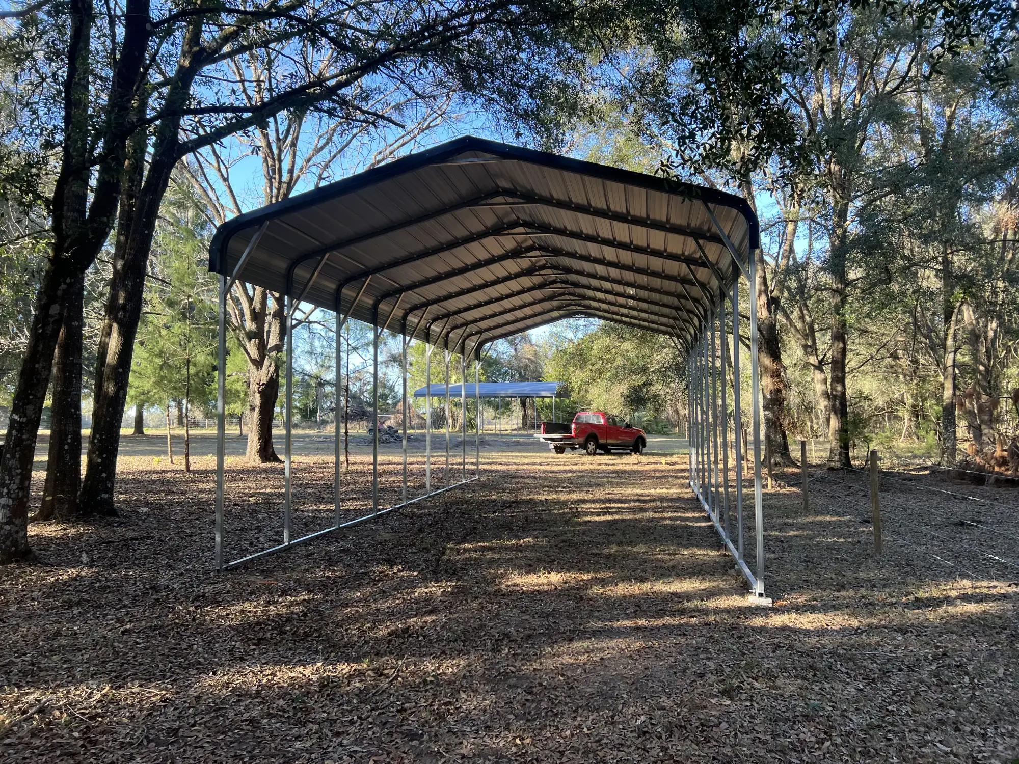 A tall open metal carport installed on a dirt driveway surrounded by trees in Ocala, FL, with a red pickup truck parked behind it.