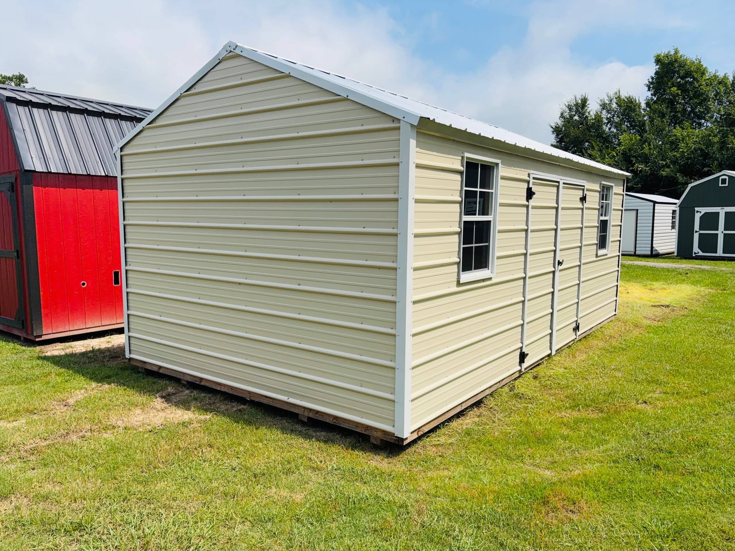 Light beige metal storage building featuring white trim, side double doors, two grid windows, and a metal gabled roof sitting on a grassy lot in Williston, FL.