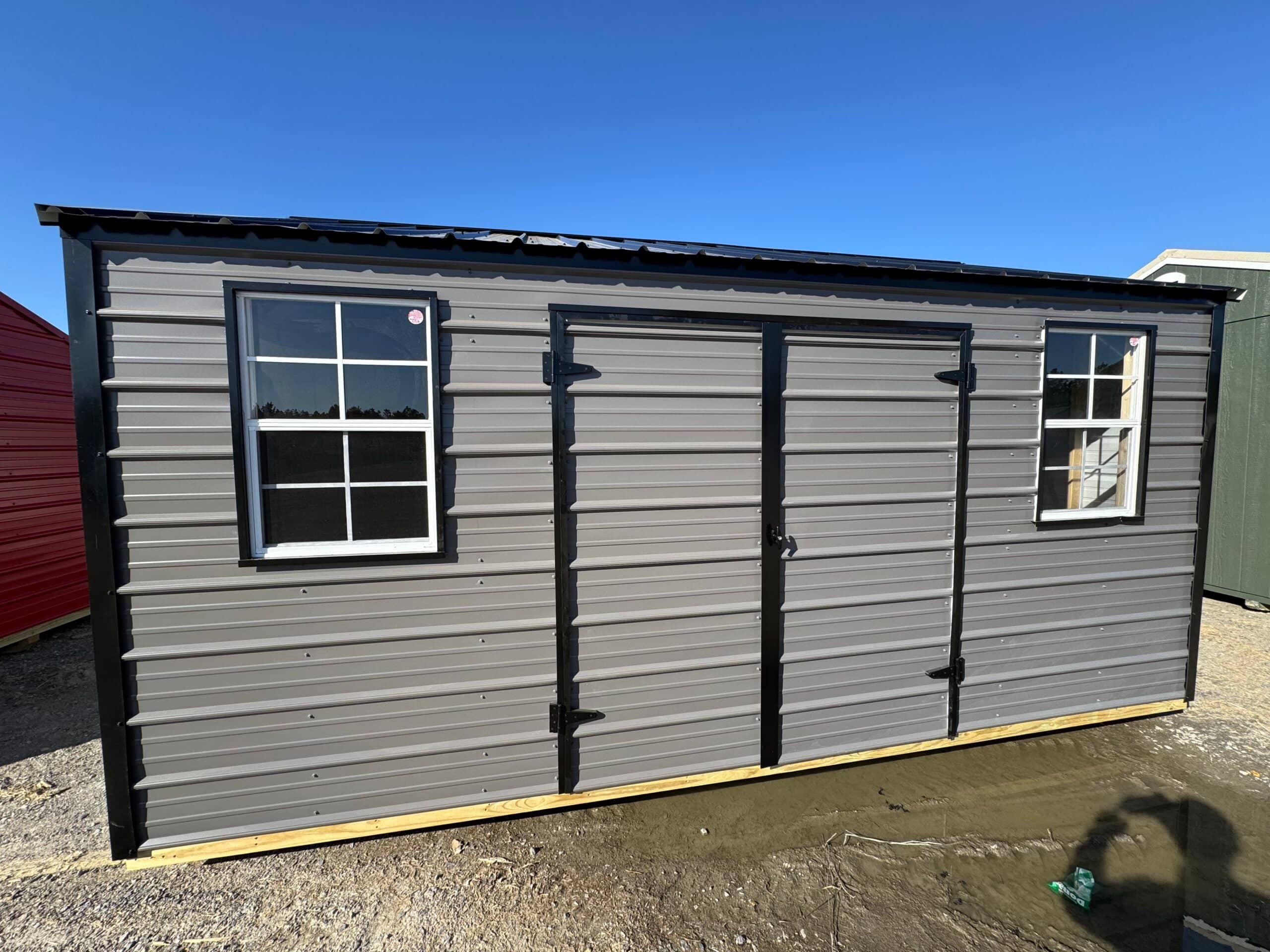 Exterior front view of a grey metal side utility shed with black trim, central double doors, and two grid windows on a gravel surface in Inverness, FL.