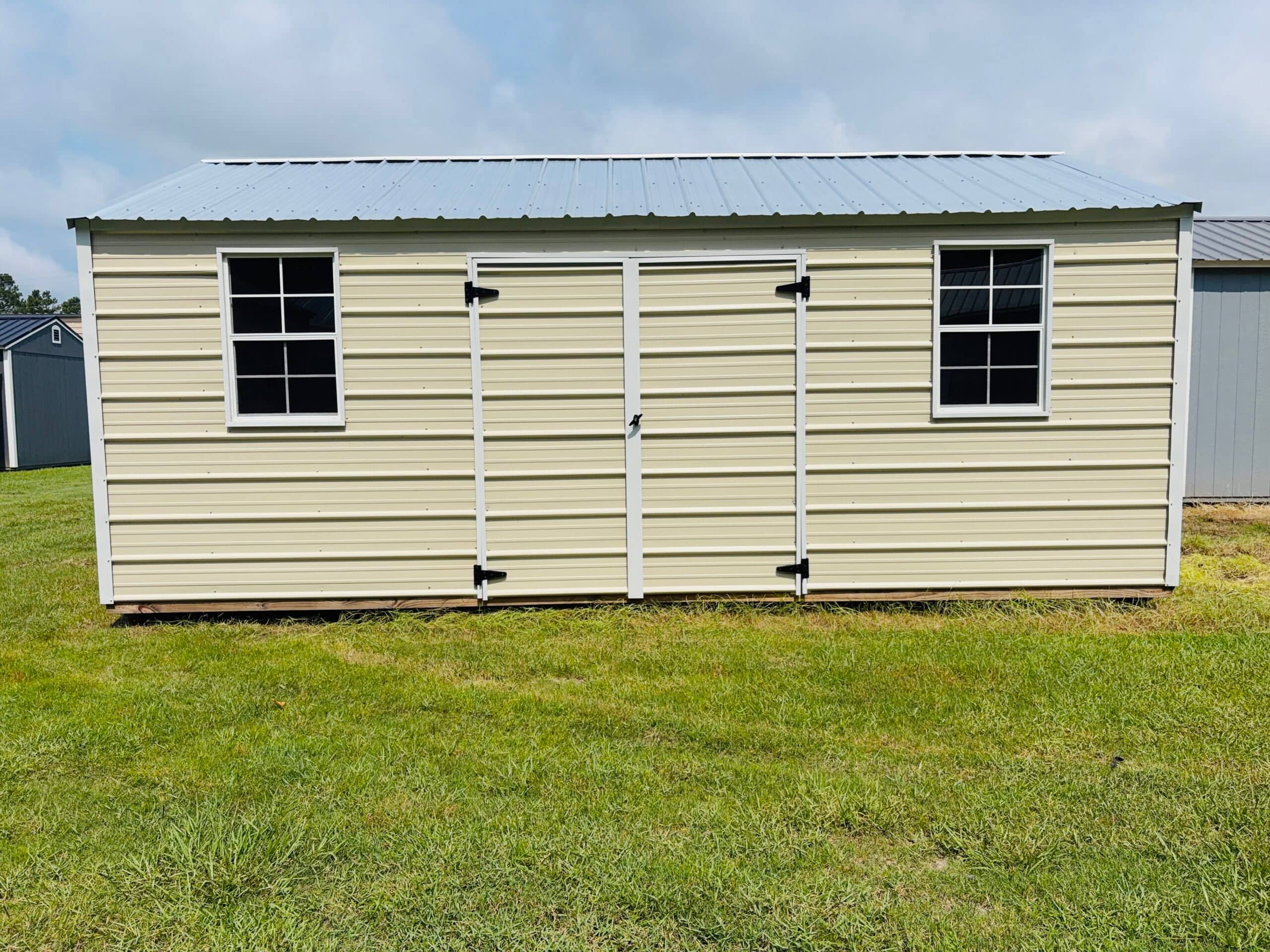 Light cream metal utility storage shed featuring white trim, double center doors, two grid windows, and a silver metal roof sitting on a grassy yard in Gainesville, FL.