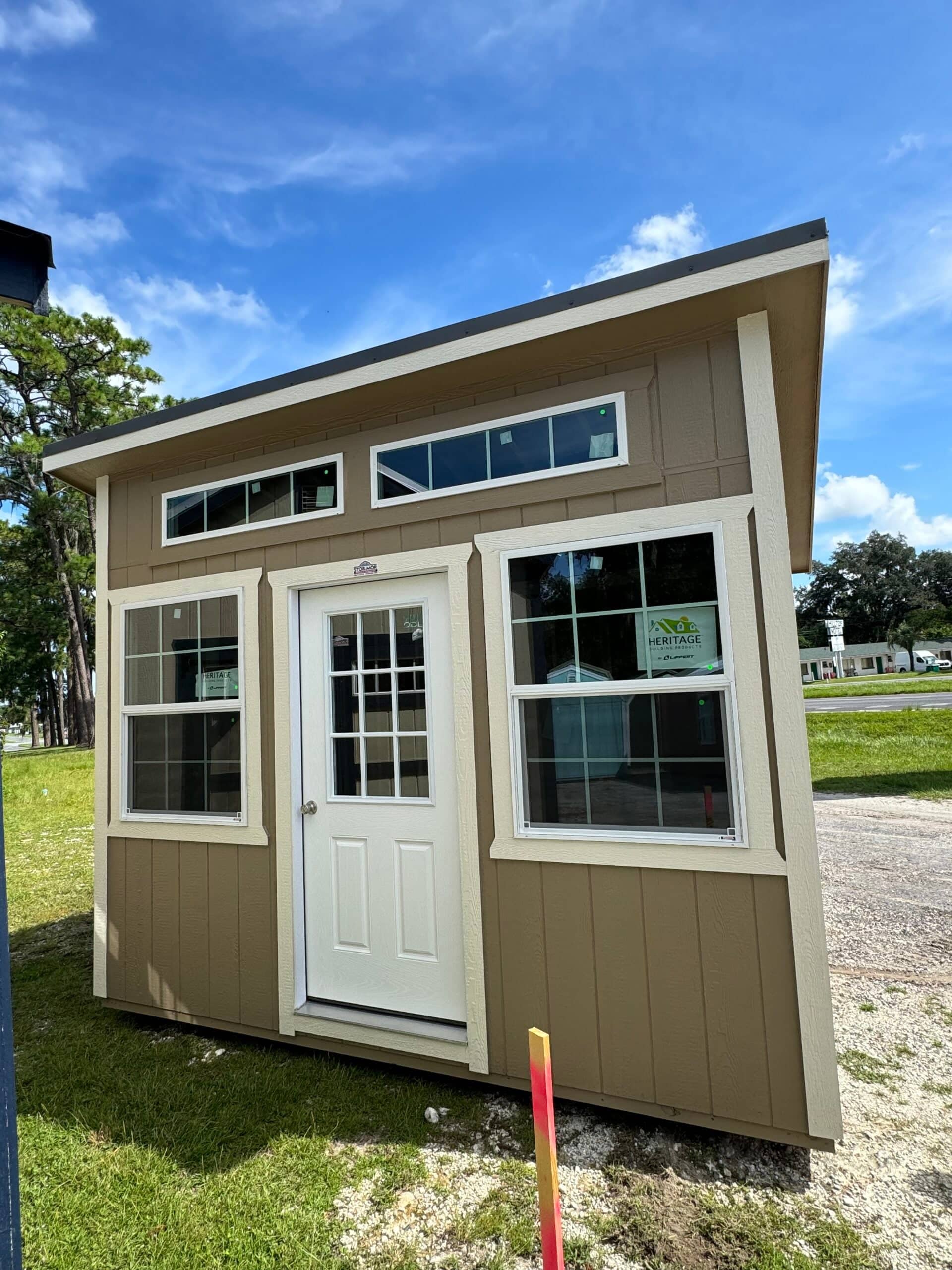 Exterior view of a tan modern sloped roof studio shed featuring beige trim, upper transom windows, grid side windows, and a glass-paneled white door in Inverness, FL.