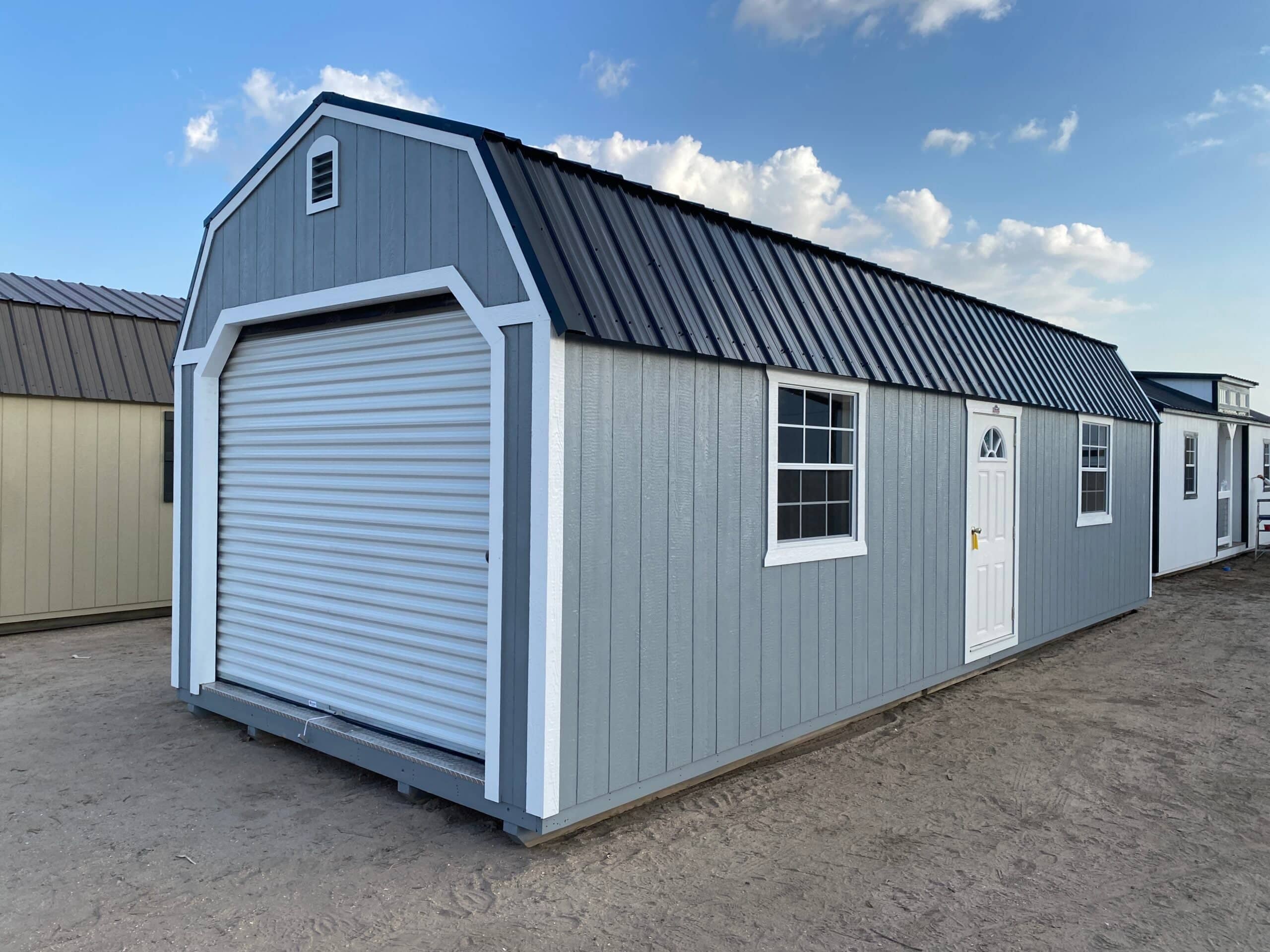 Exterior view of a grey high-barn garage shed with white trim, a metal roof, roll-up front garage door, side house door, and two grid windows in Brooksville, FL.